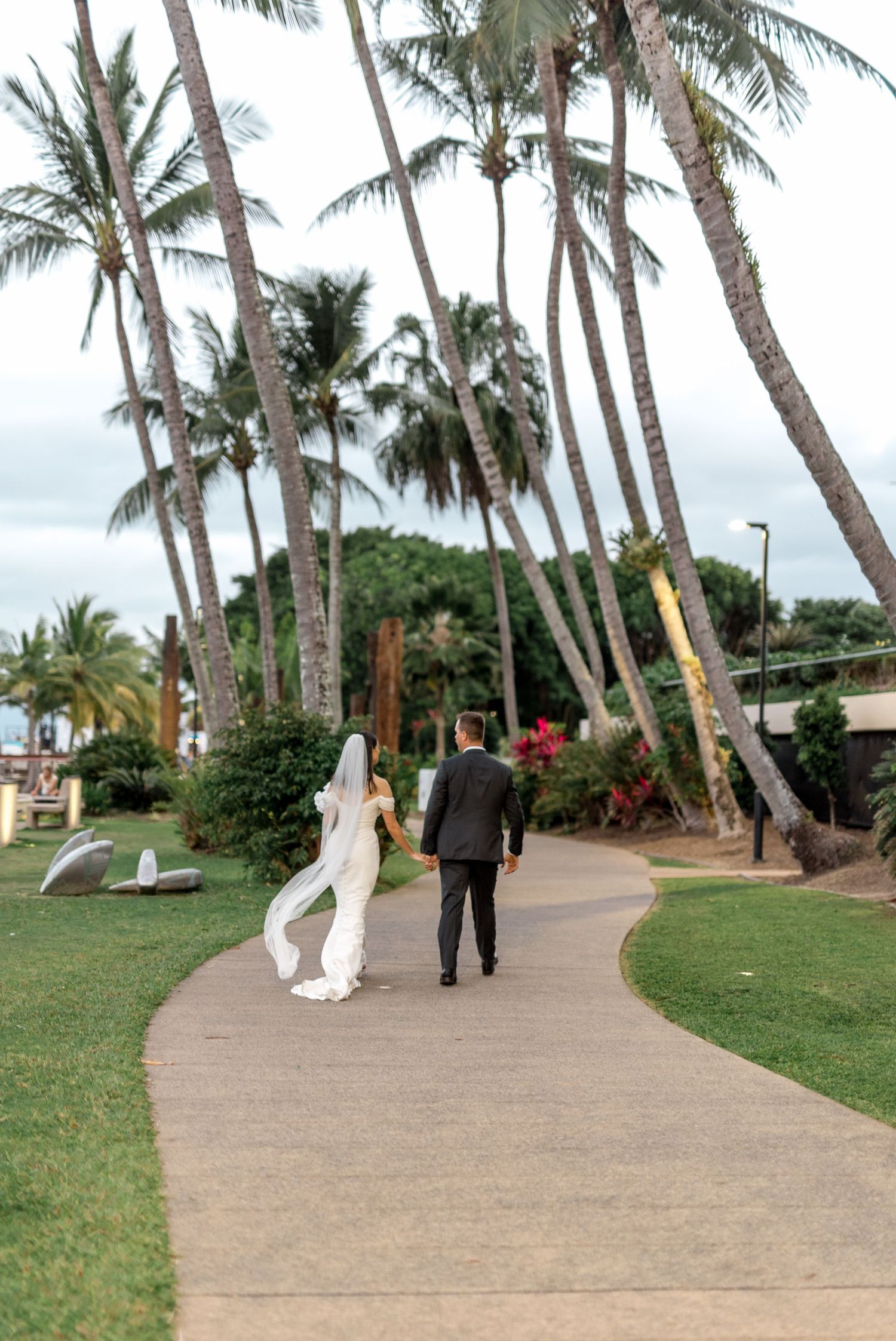 Bride and groom walking away on a curved path surrounded by tall tropical palm trees at a Hawaiian wedding venue.