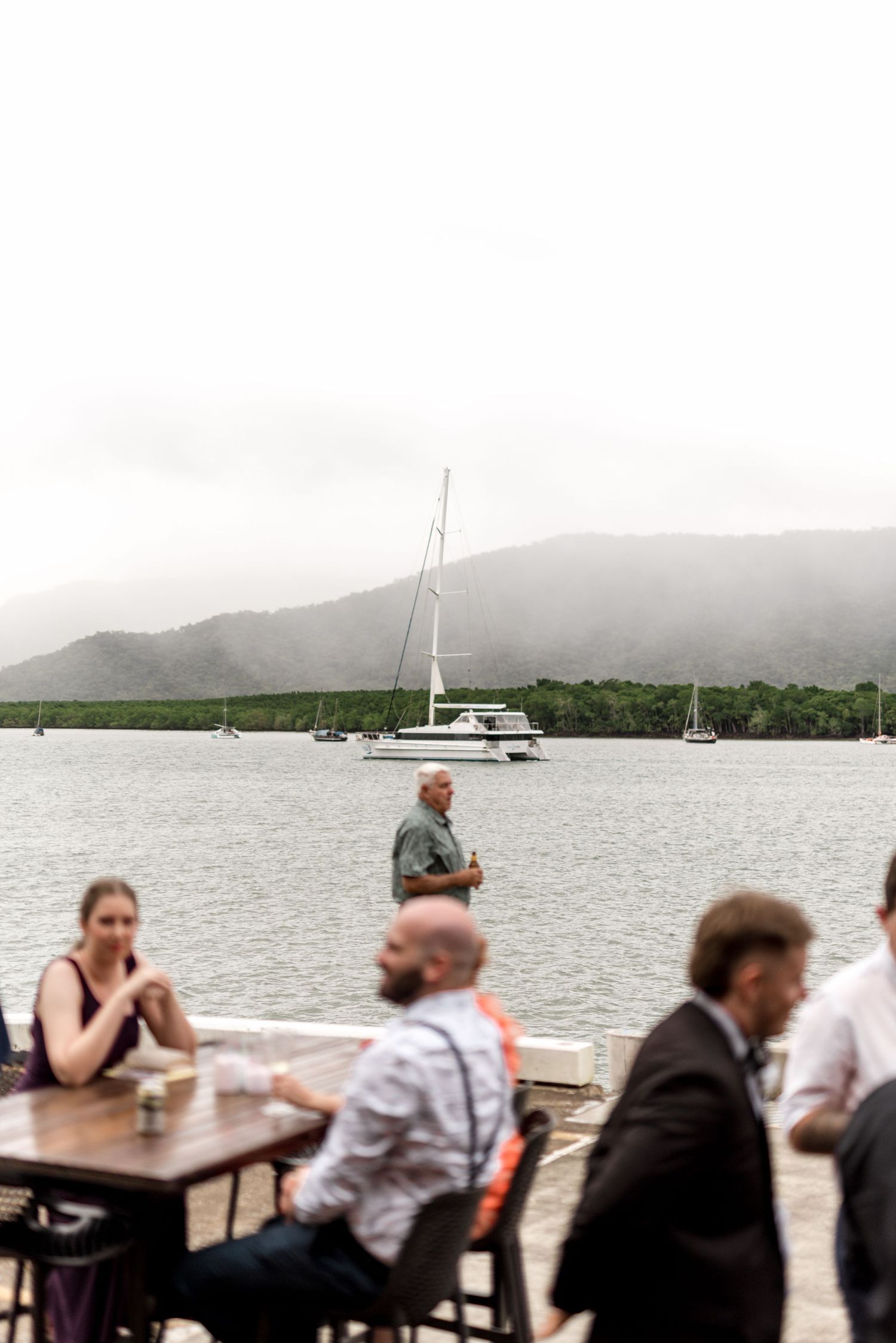 Wedding guests seated and standing at a waterfront reception with sailboats and misty mountains in background.