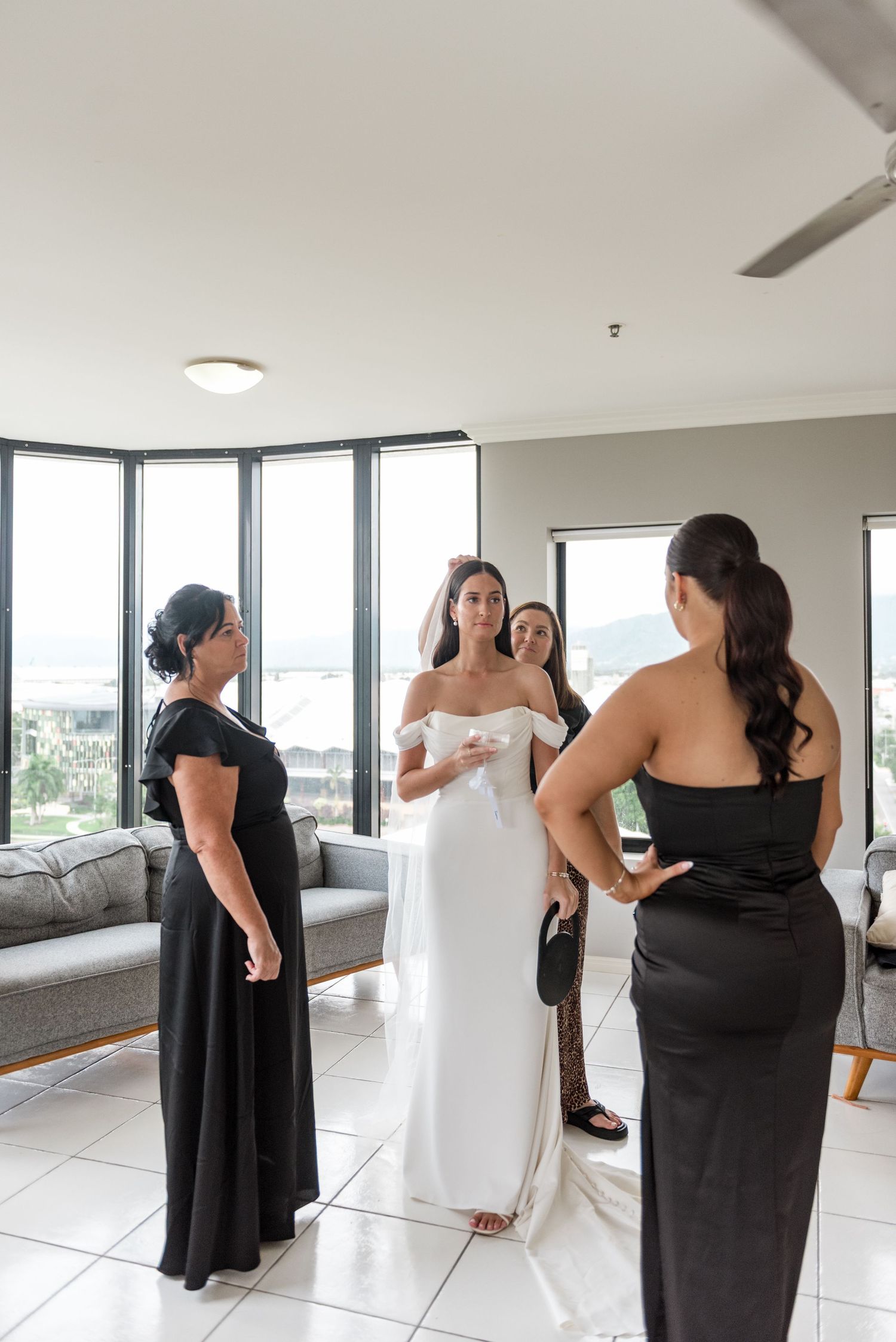 Bride in white off-shoulder gown stands with bridesmaids in black dresses in a modern apartment before wedding.