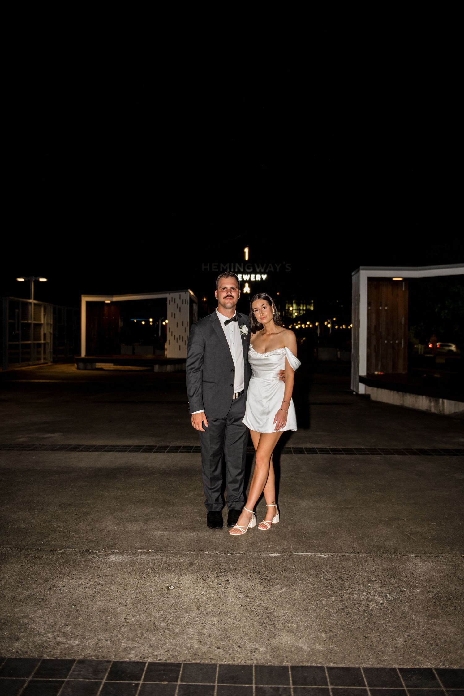Bride in white mini dress and groom in dark suit pose together outside at night.