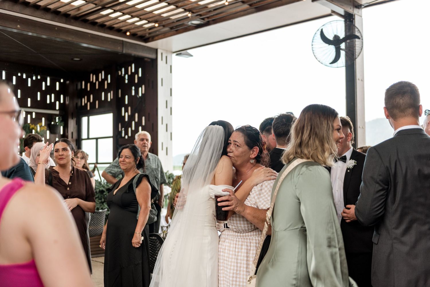 Bride in white veil embracing female guest warmly during wedding reception celebrations.