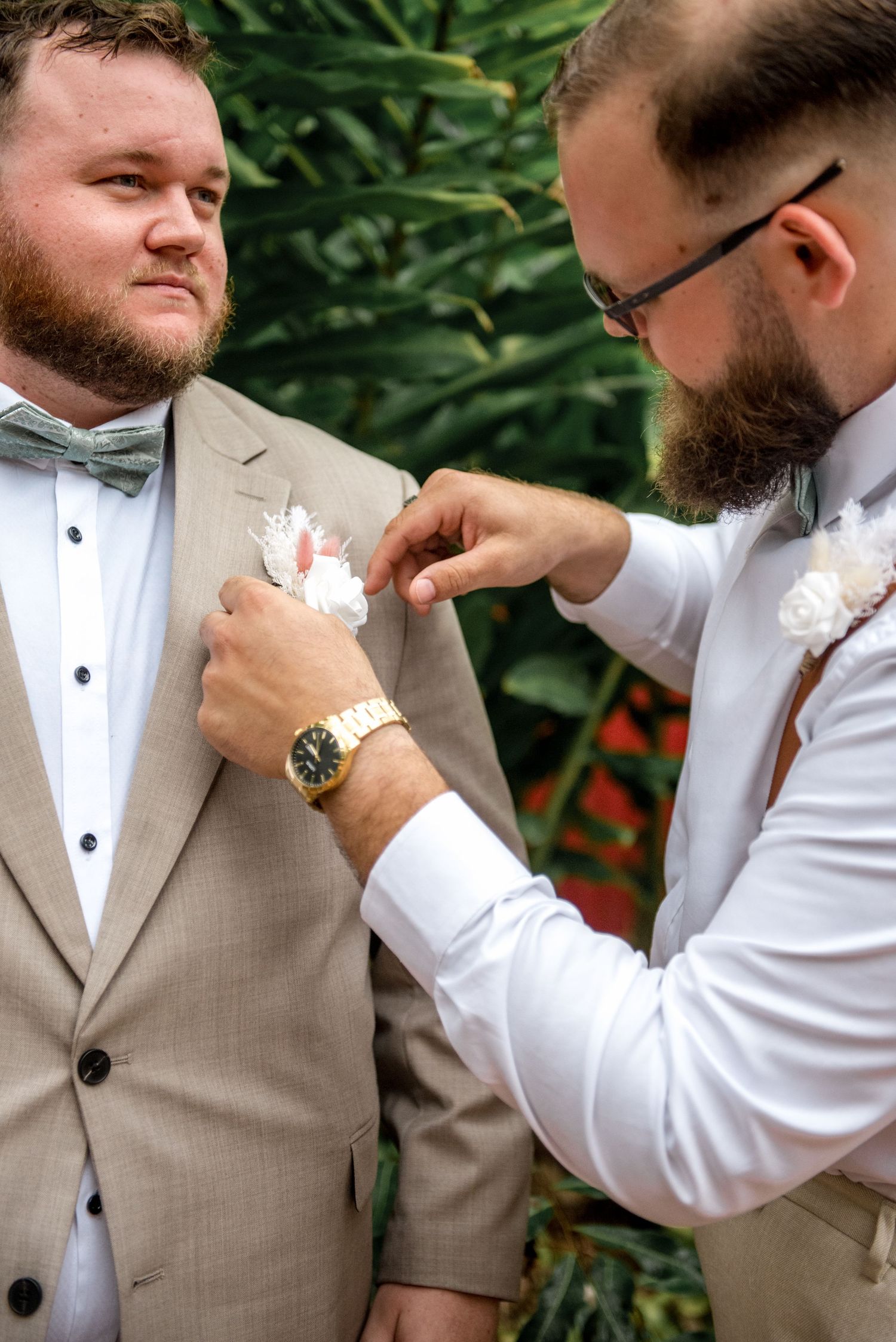Groomsman pinning a white boutonniere on the groom's tan suit lapel at a wedding.