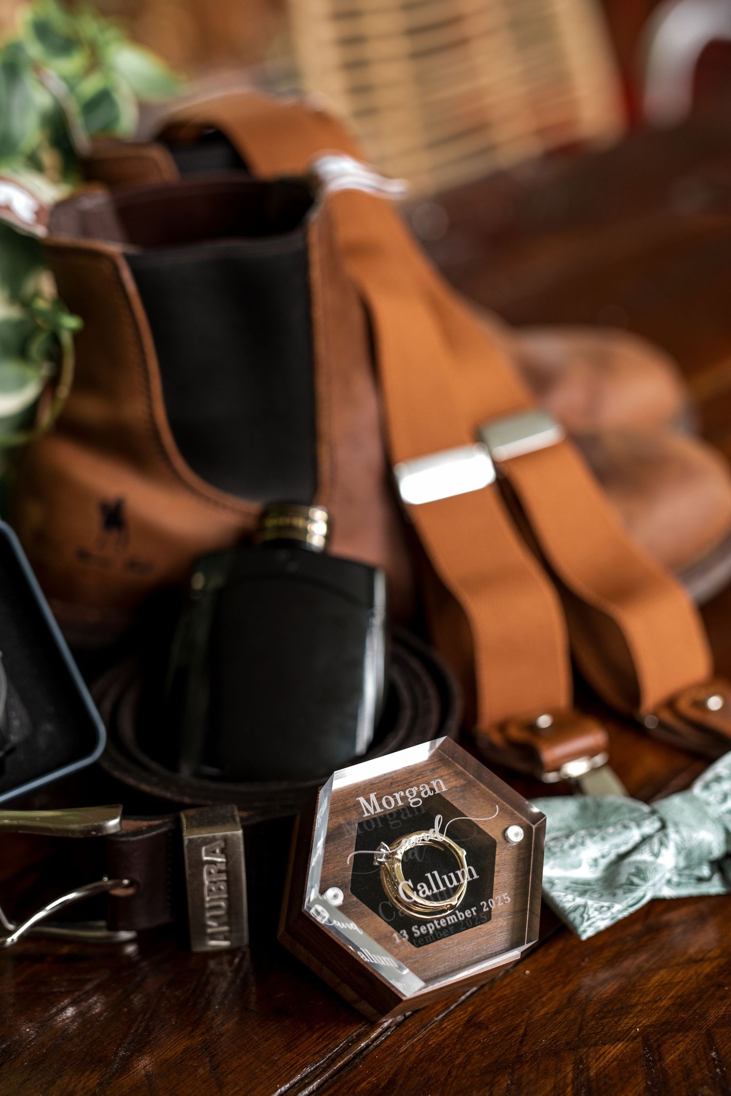 Groom's wedding flat lay featuring leather suspenders, flask, belt, bow tie, and custom engraved ring box.