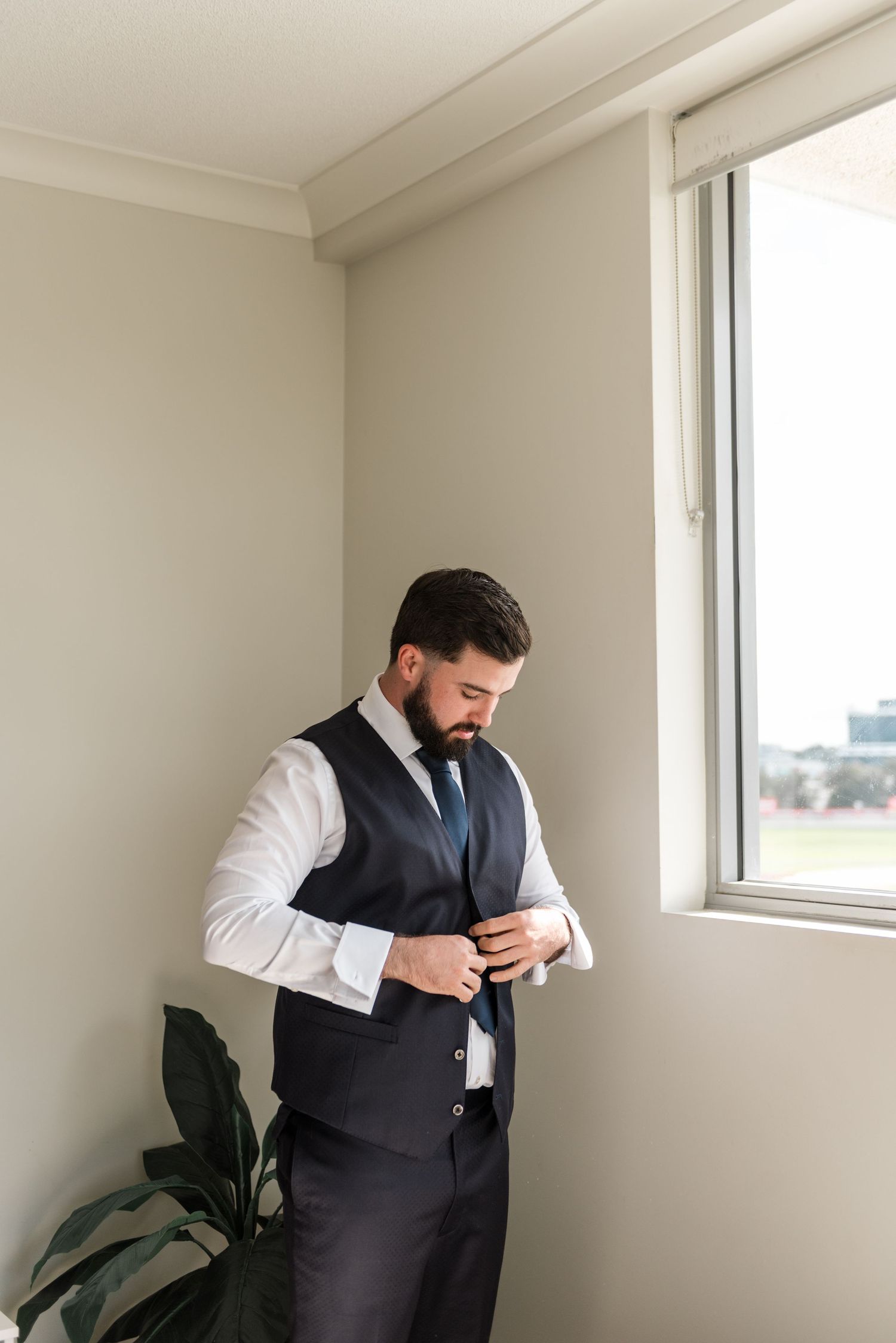 Groom in a navy vest and white shirt fastening his cufflinks by a sunlit window.