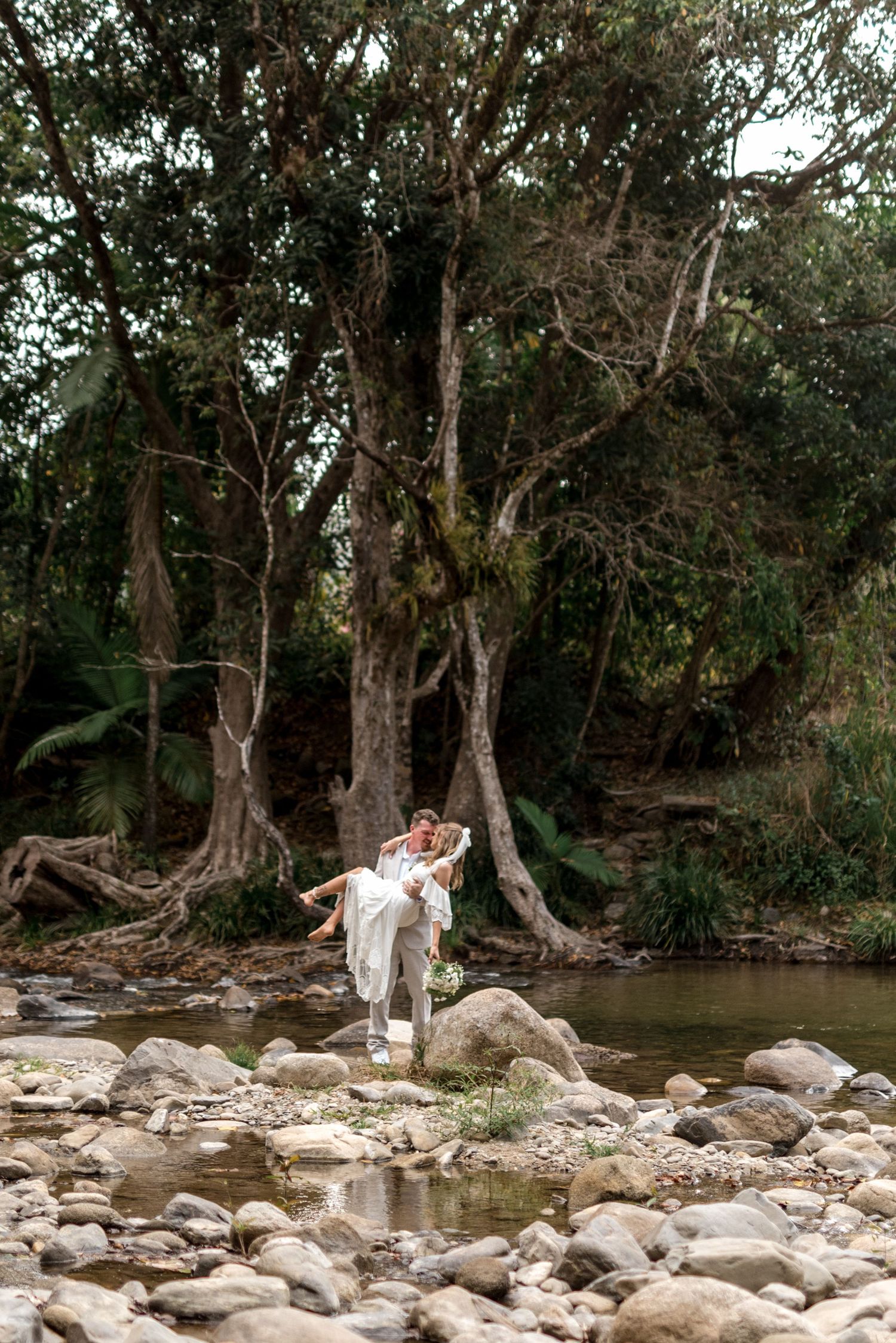 Groom carries bride in his arms among large trees and rocky creek in a lush forest setting.