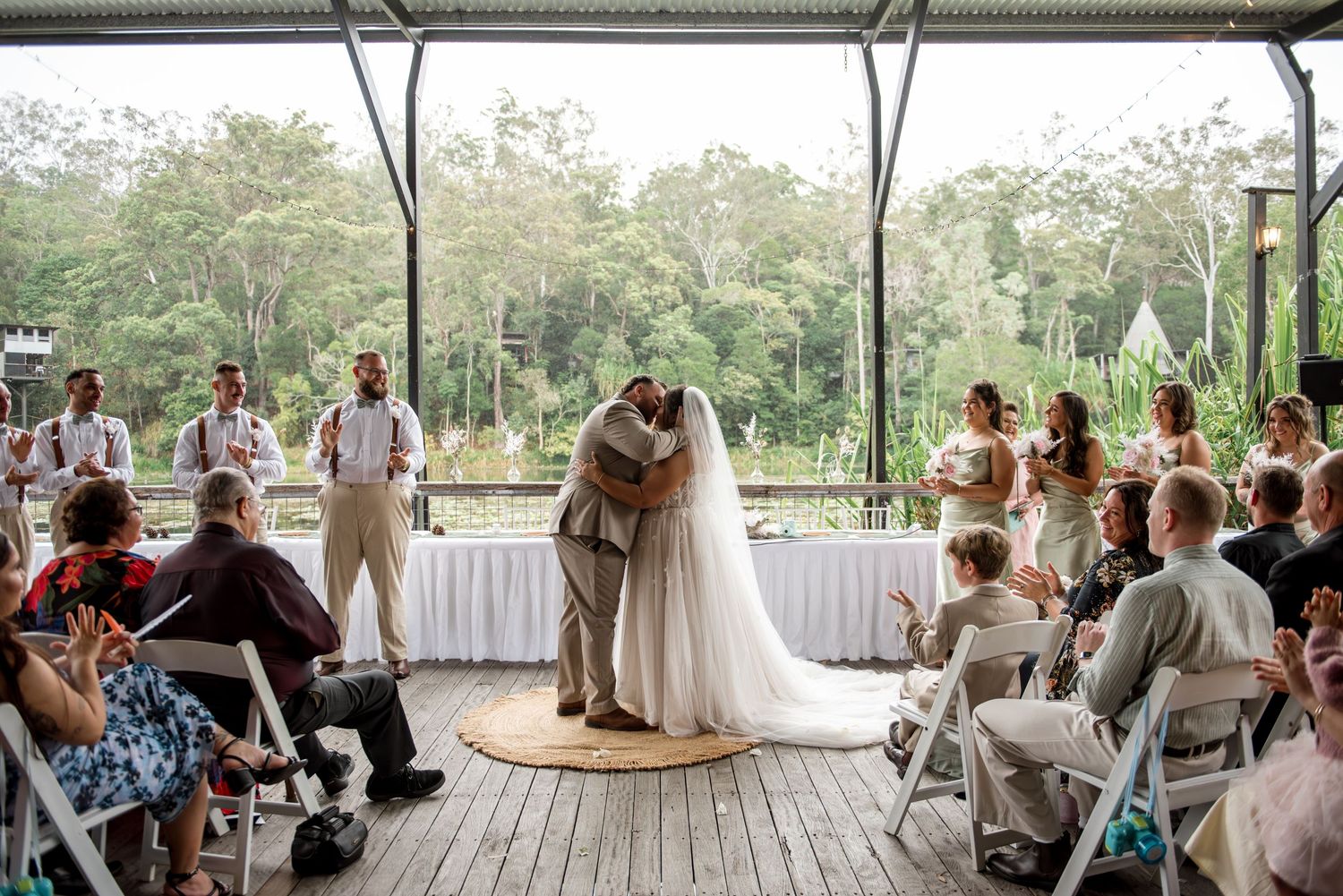 Bride and groom share first kiss surrounded by wedding party and seated guests in outdoor venue.