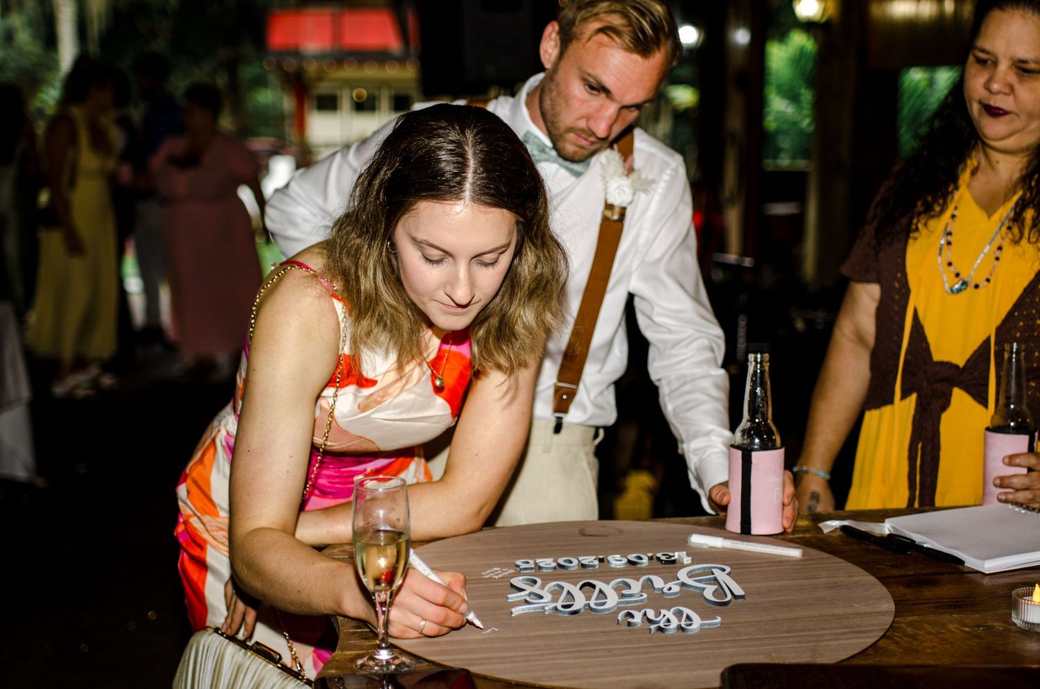 Wedding guest in colorful dress signs guestbook while holding champagne glass at reception table.