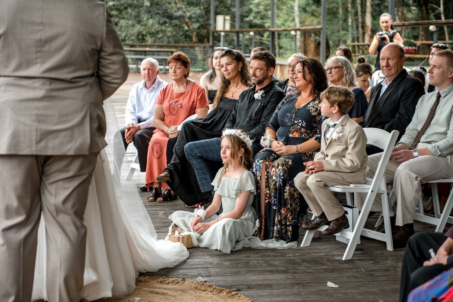Wedding guests smile watching bride walk down aisle with flower girl seated nearby.