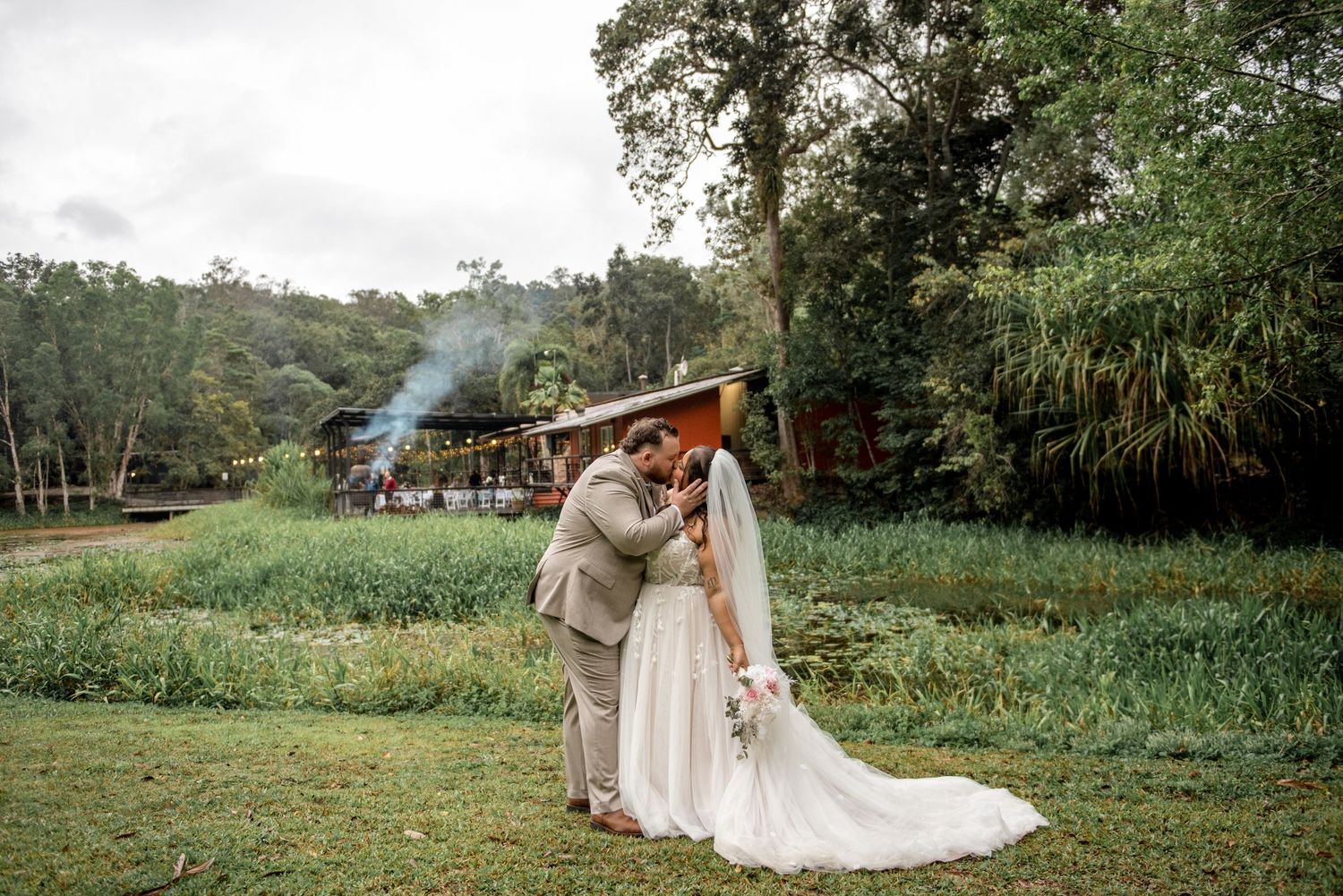 Bride and groom share a tender kiss in a lush green garden with a rustic venue backdrop.