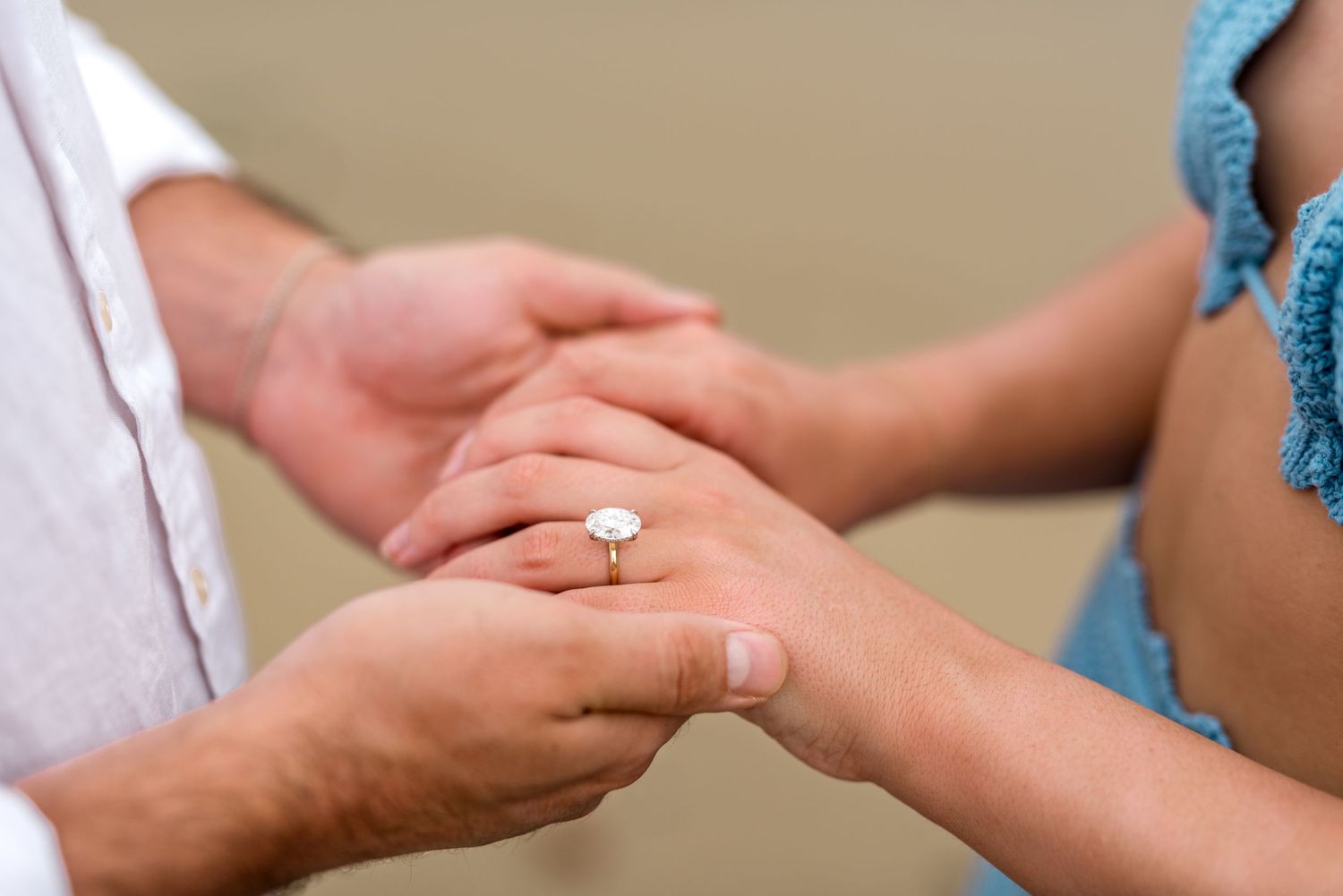 Close-up of an engagement ring on a woman's finger as a man holds her hand on a beach.