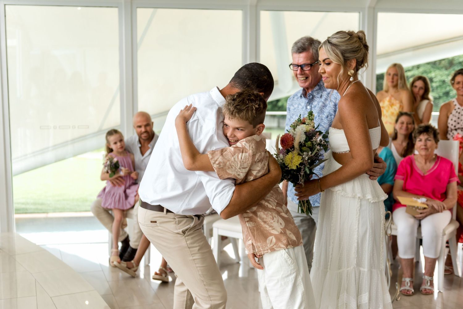 Groom bending down to hug a child beside the bride during wedding ceremony.