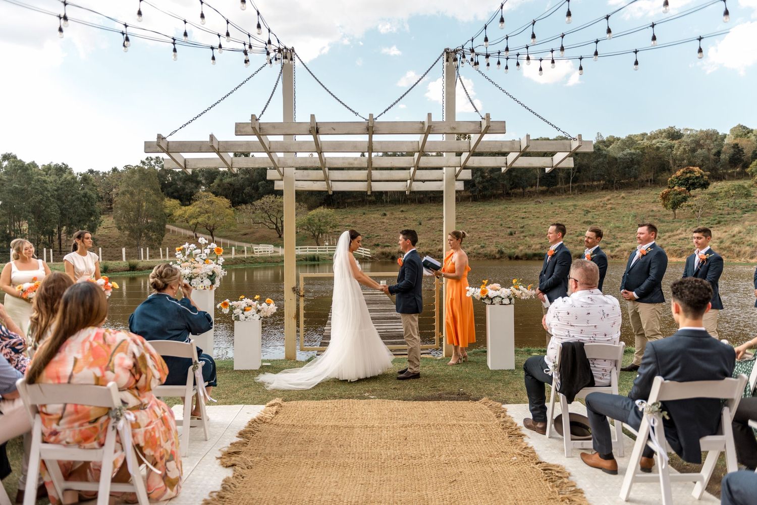 Bride and groom exchange vows under a white pergola by a pond, with bridesmaids and groomsmen standing nearby.