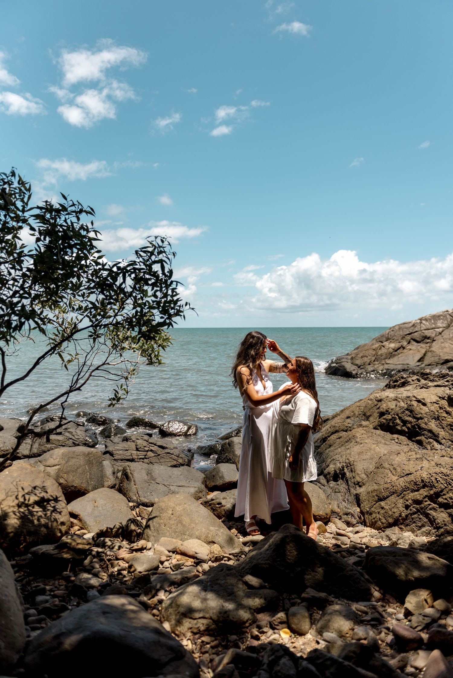 Two women in white dresses share a tender moment on rocky shoreline with blue ocean and cloudy sky behind them.