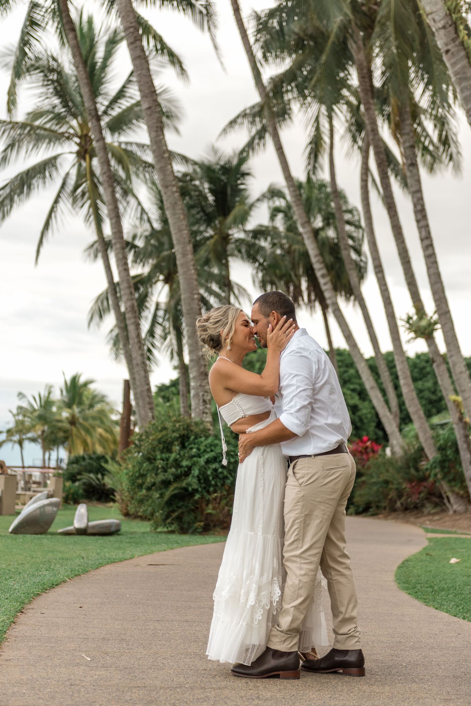 Couple sharing a romantic kiss on a garden path surrounded by tall palm trees at a tropical wedding venue.