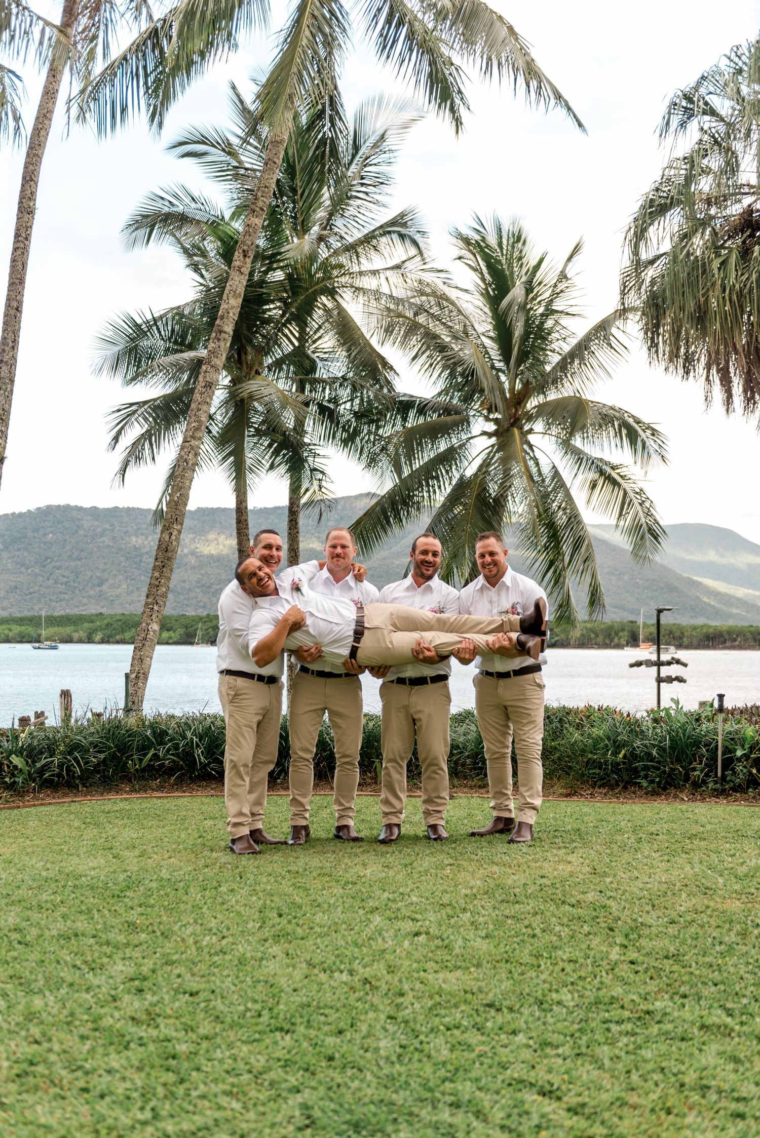Three groomsmen playfully lift the groom horizontally near palm trees by a tropical waterway.