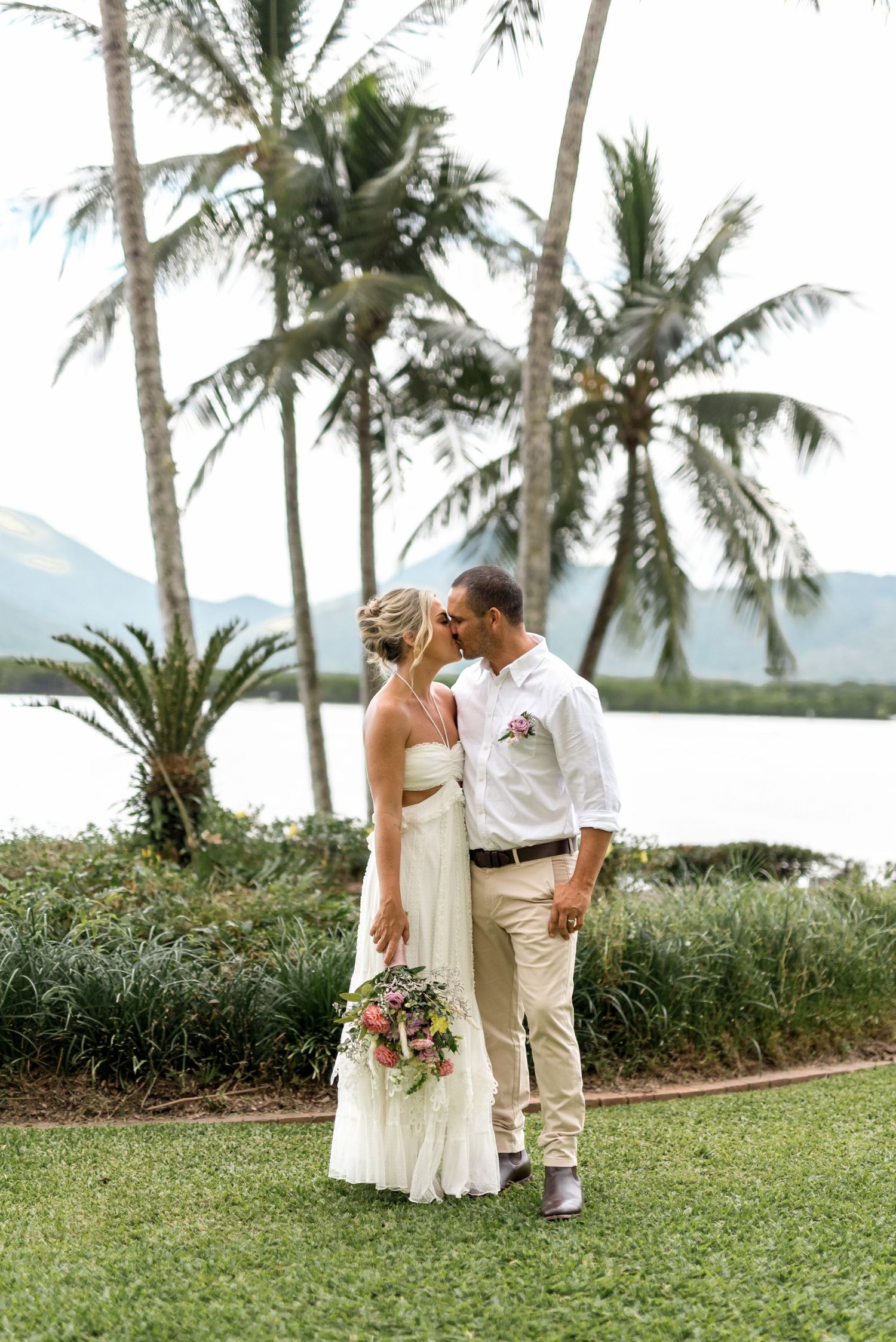 Newlyweds share a tender kiss on lush lawn surrounded by tall palm trees and serene waterfront.