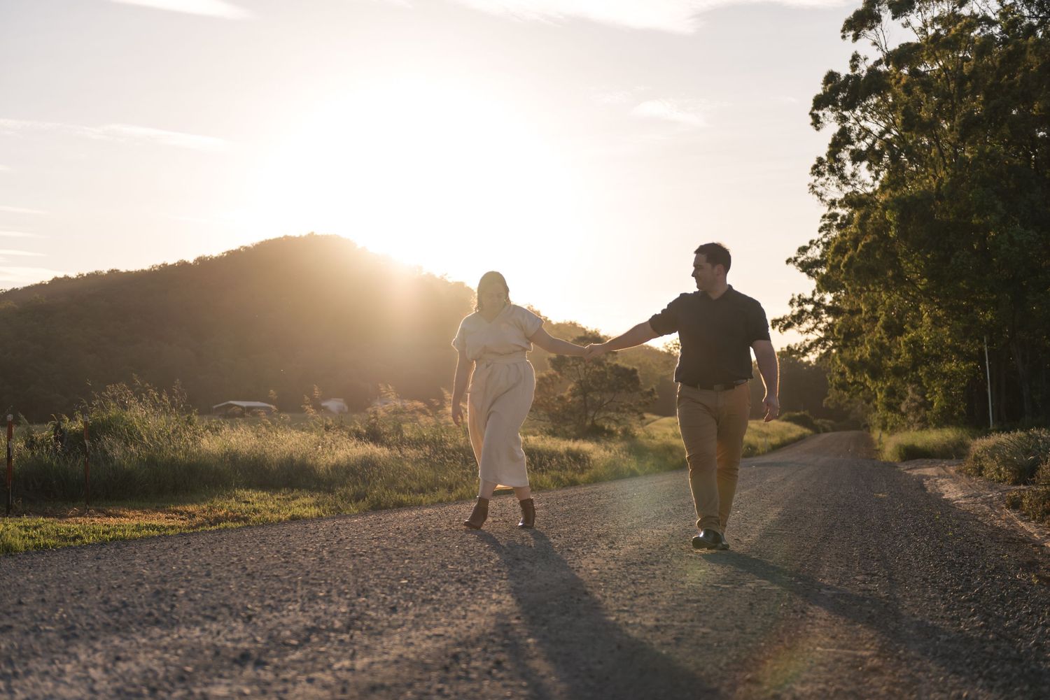 Couple holding hands strolling along country road with warm sunset glow and trees.