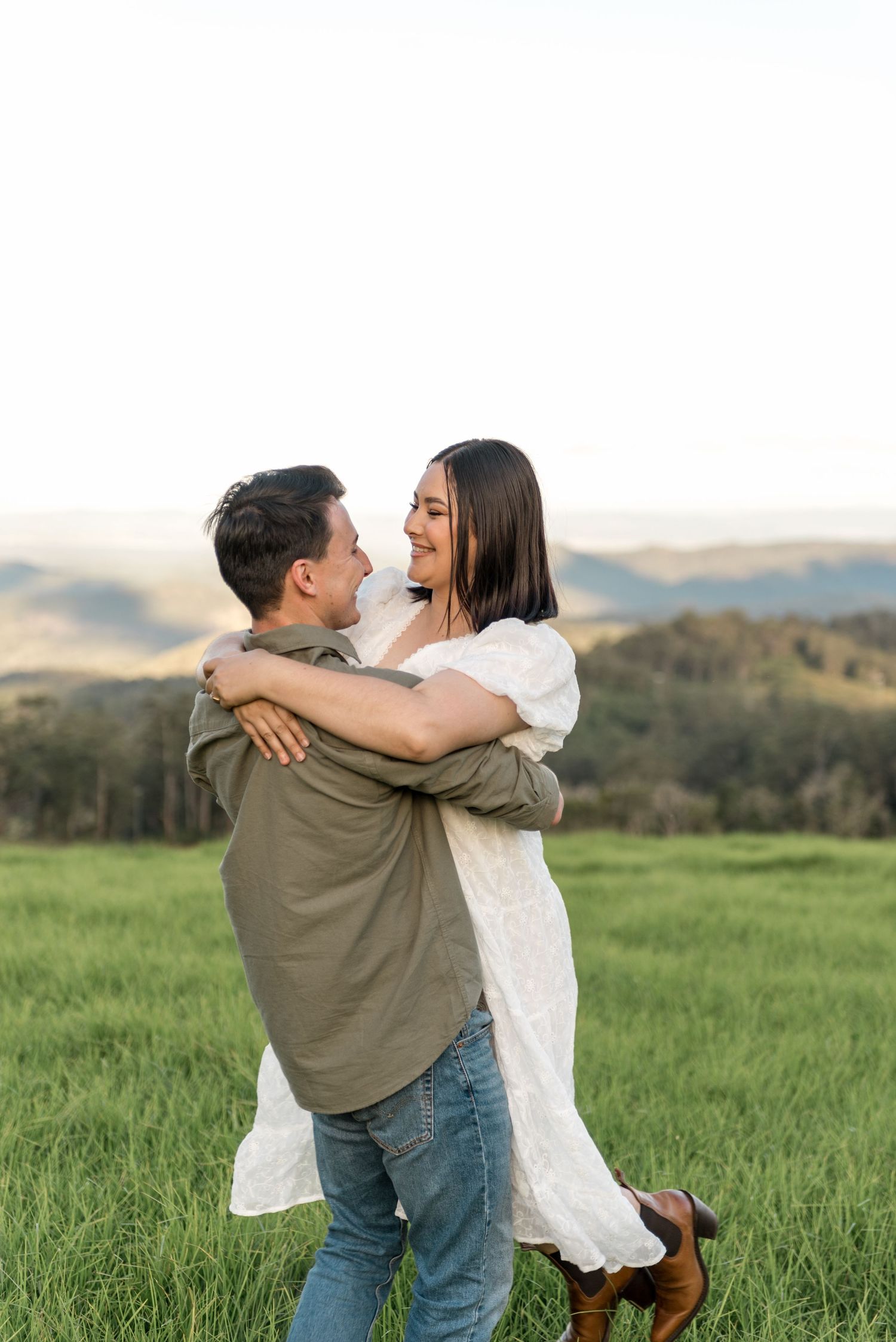 Man lifting smiling woman in a green field with rolling hills at sunset.