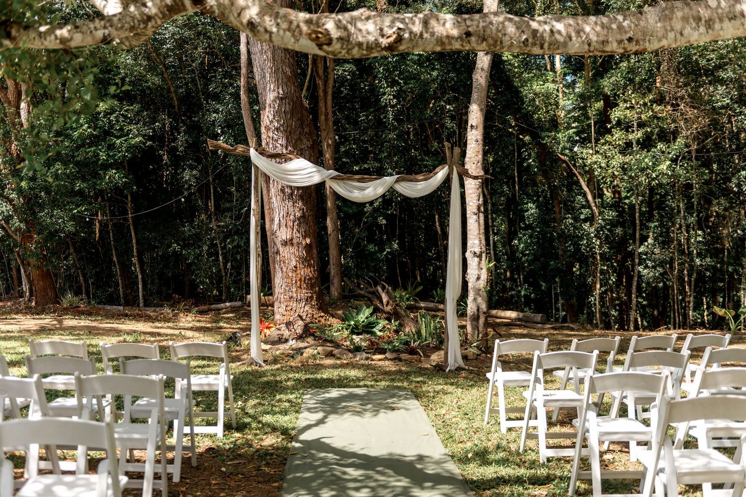 Rustic outdoor wedding ceremony with white chairs and fabric-draped tree arch in a lush forest.