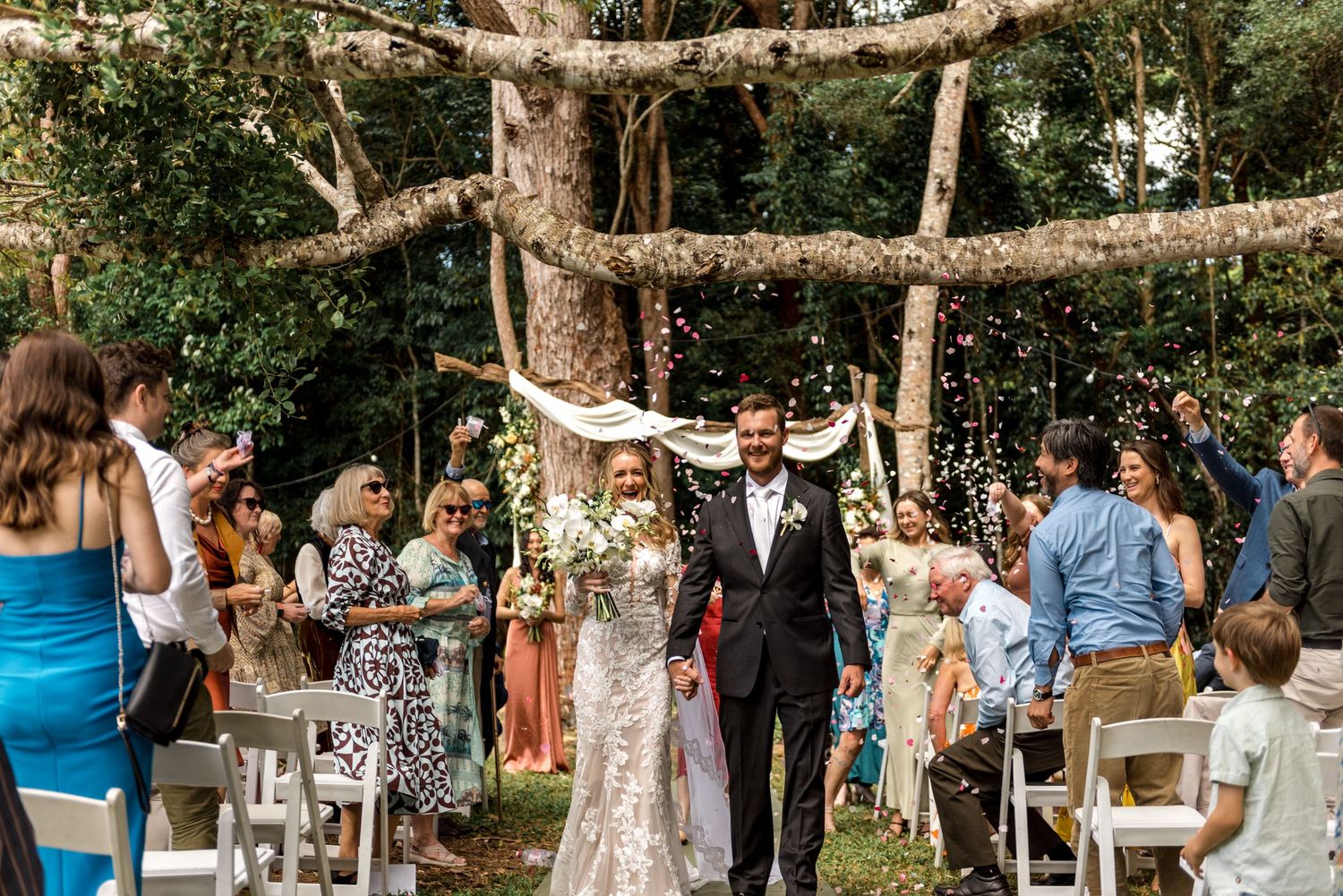 Newlyweds walk down aisle as guests celebrate with confetti in forest setting.