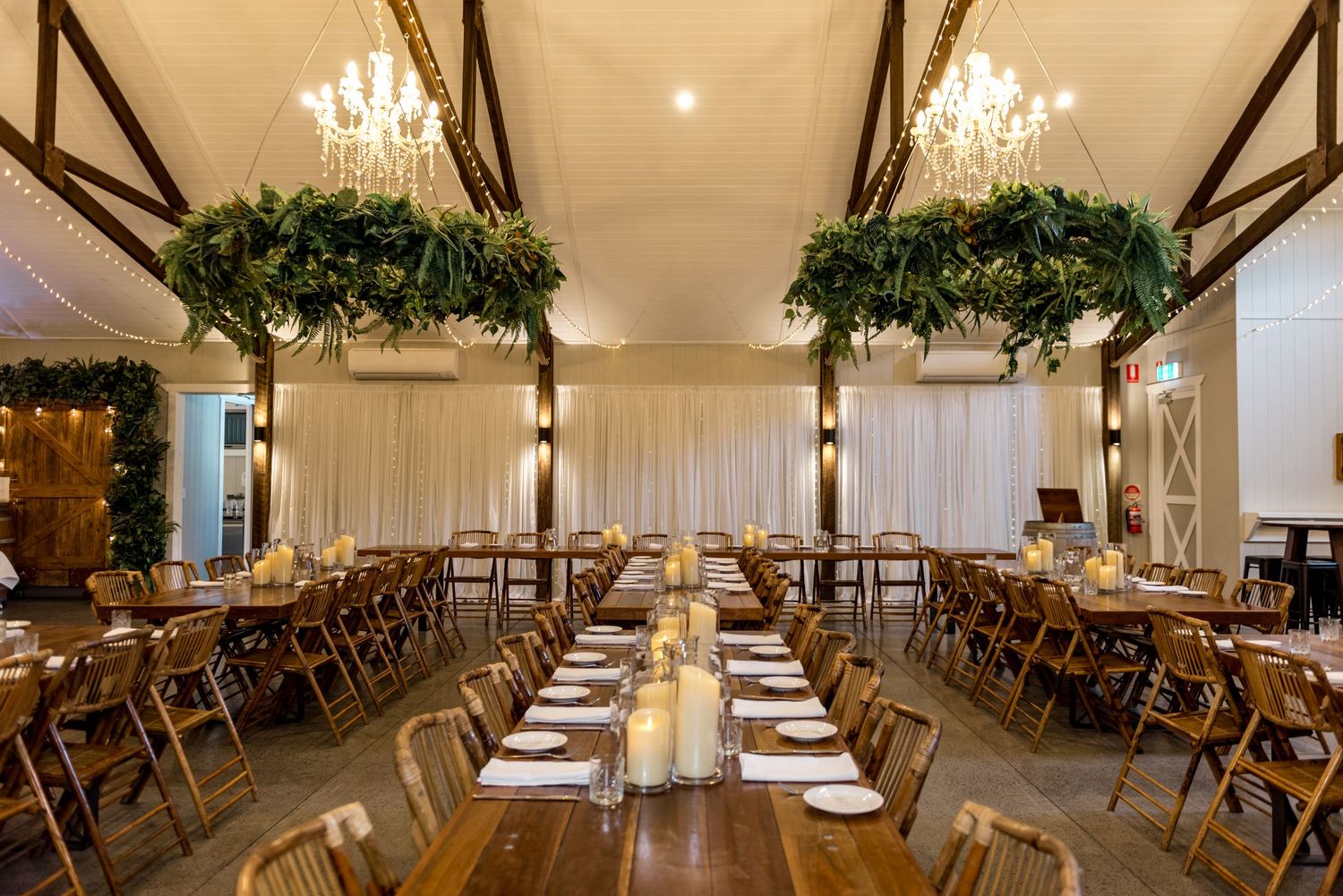 Elegant wedding reception barn with long wooden tables, fairy lights, chandeliers, and lush green floral installations.