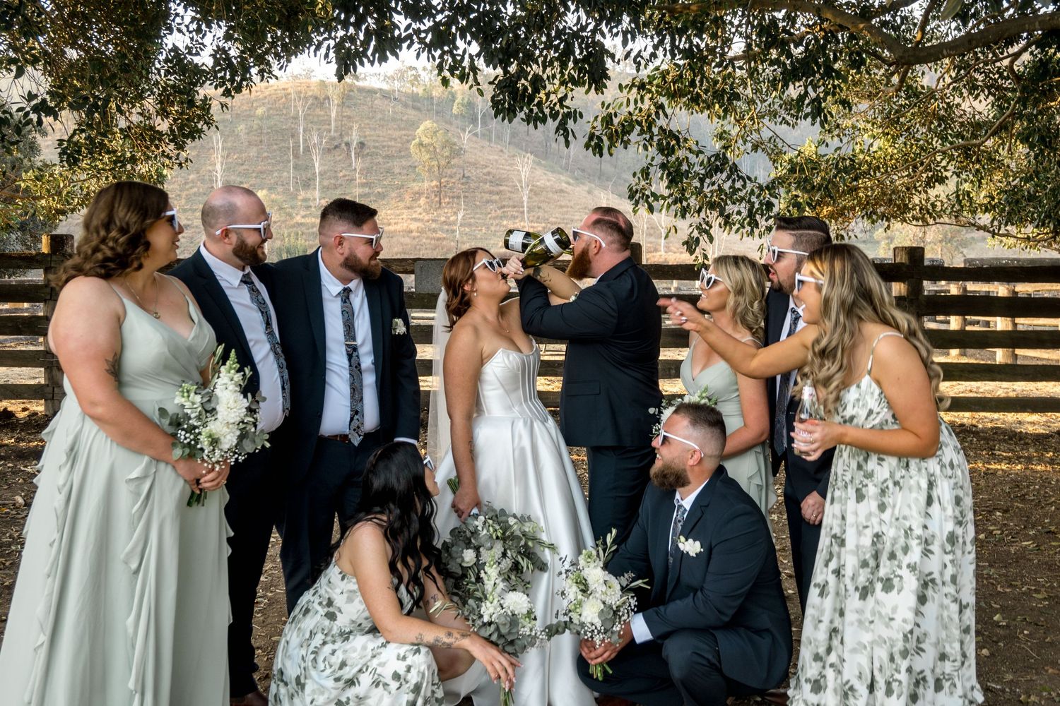 Wedding party laughing and celebrating together outdoors near rustic wooden fence at farm venue.