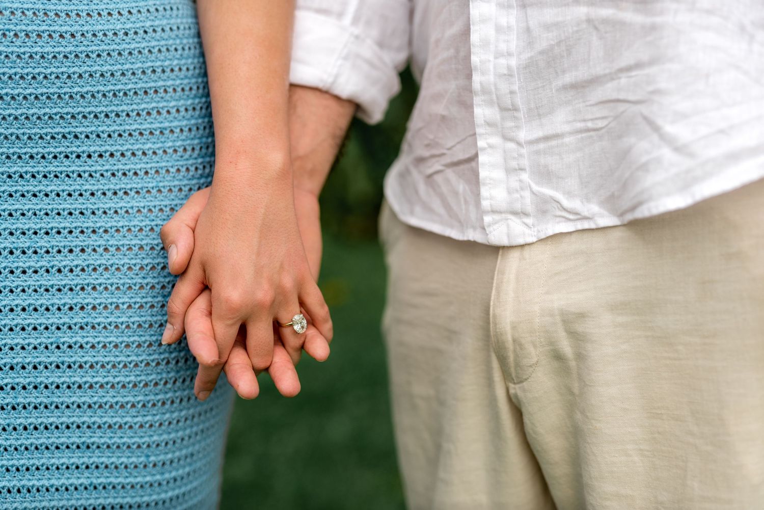 Close-up of couple holding hands showing engagement ring, wearing blue dress and linen pants.