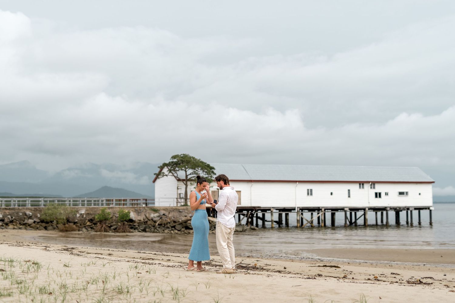 Engaged couple standing together on tropical beach near white pier building on overcast day.