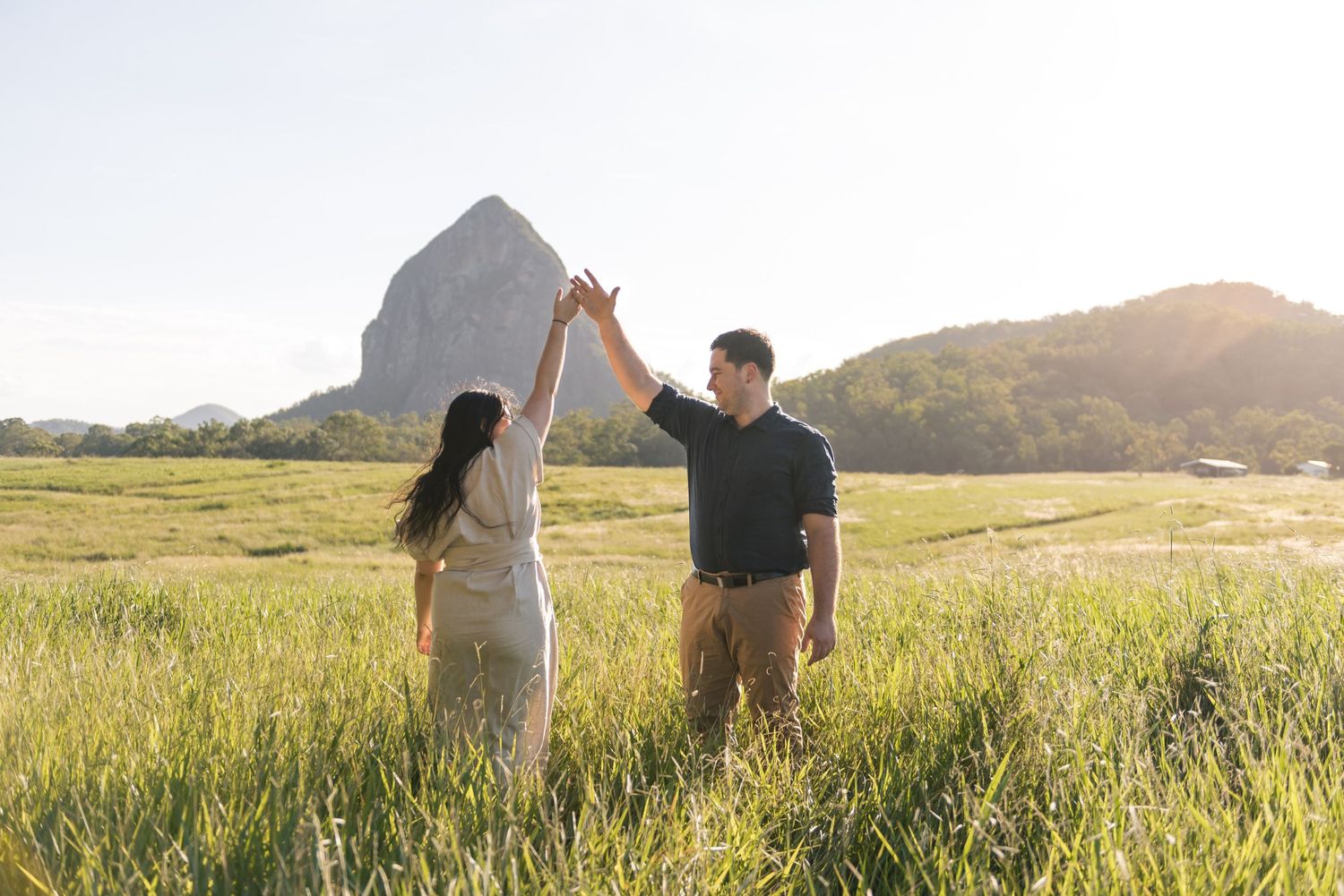 Couple holding hands and spinning in a green field with a striking rocky mountain peak at sunset.