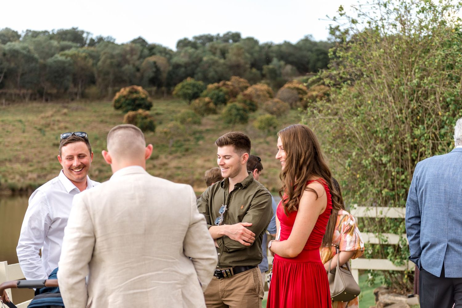 Wedding guests socializing outdoors near a white fence with lush green trees and a pond in the background.