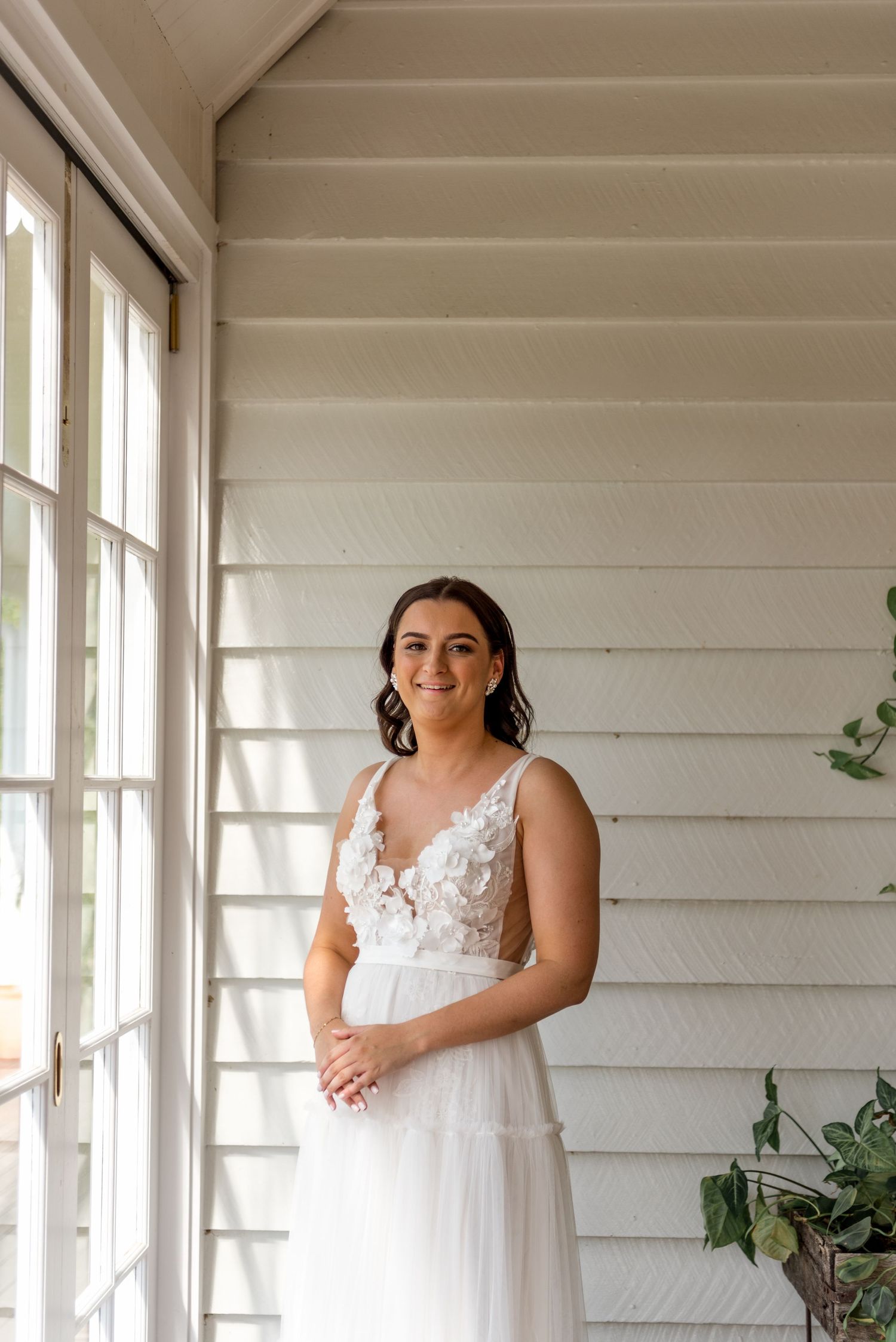 Smiling bride in white floral lace A-line wedding dress standing by window in white timber room.