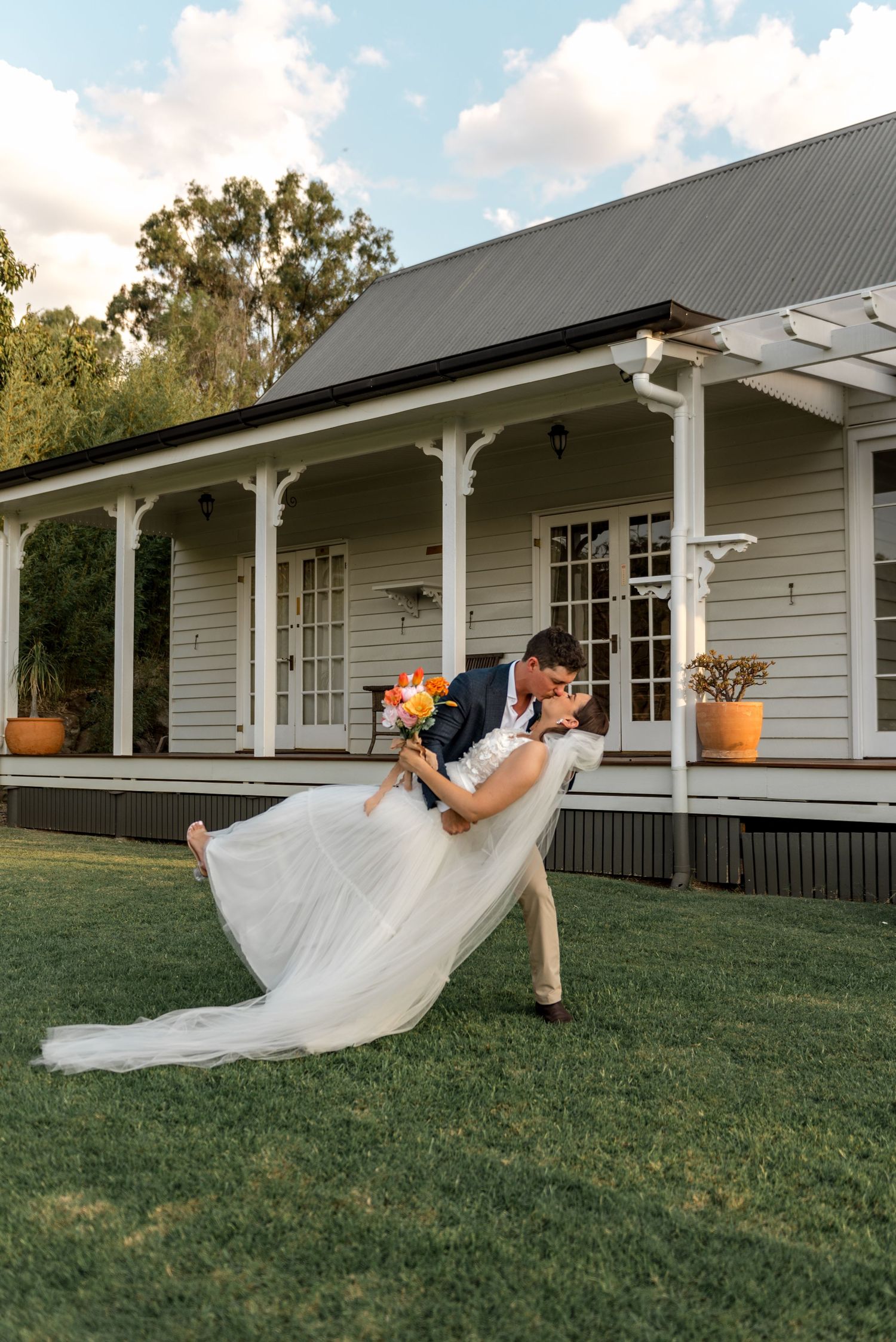 Groom dipping bride in a dramatic kiss on green lawn in front of a white farmhouse.