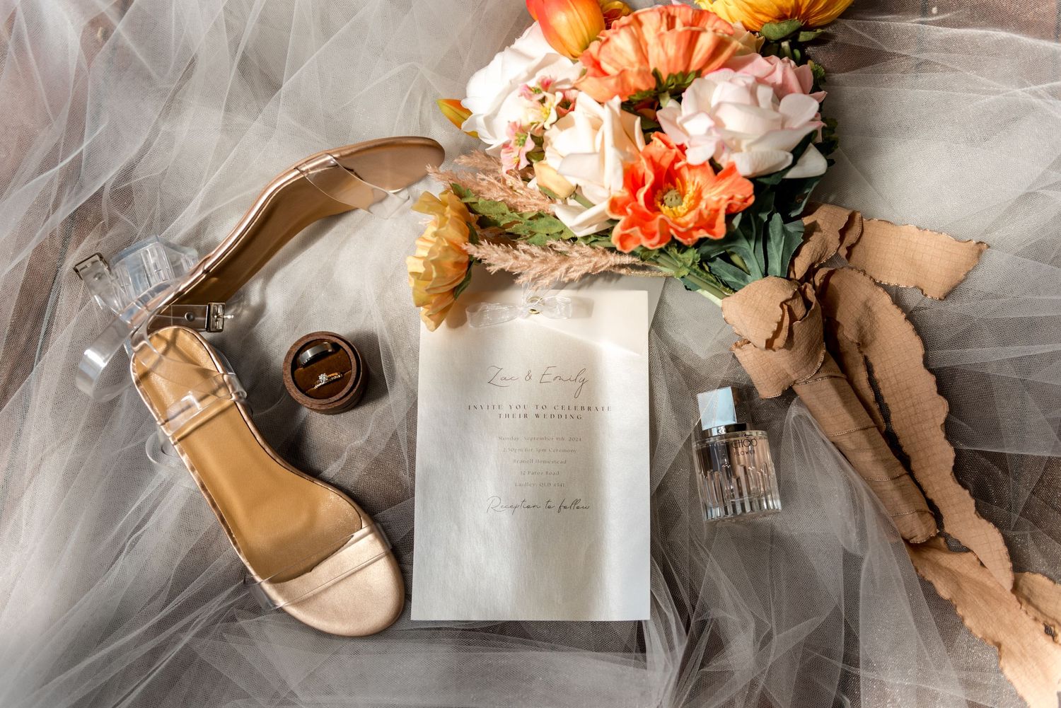 Elegant wedding flat lay with gold heels, floral bouquet, invitation, perfume, and ring on sheer veil.