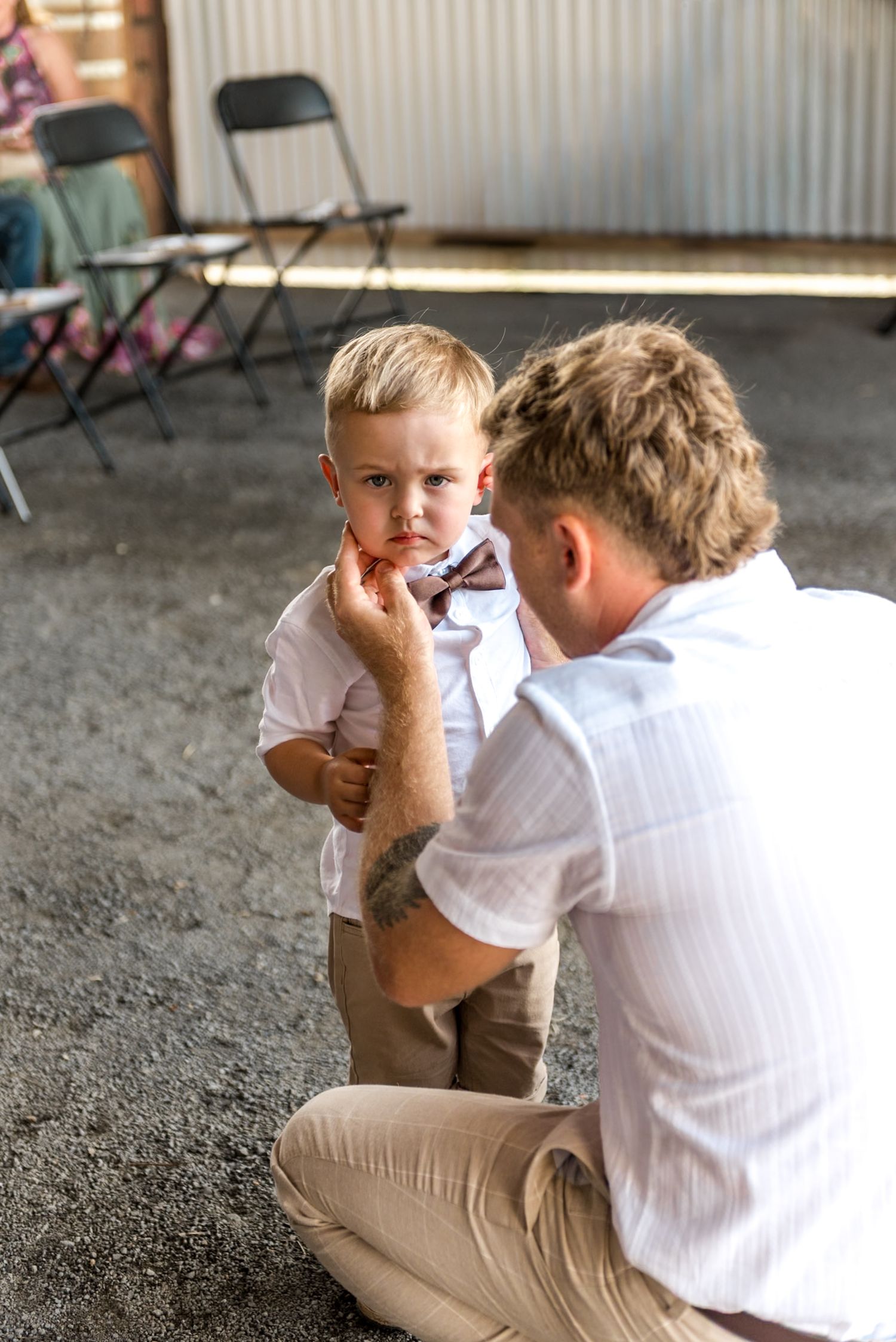 A man crouches down to adjust a bow tie on a young blonde boy in a white shirt at a rustic barn wedding.
