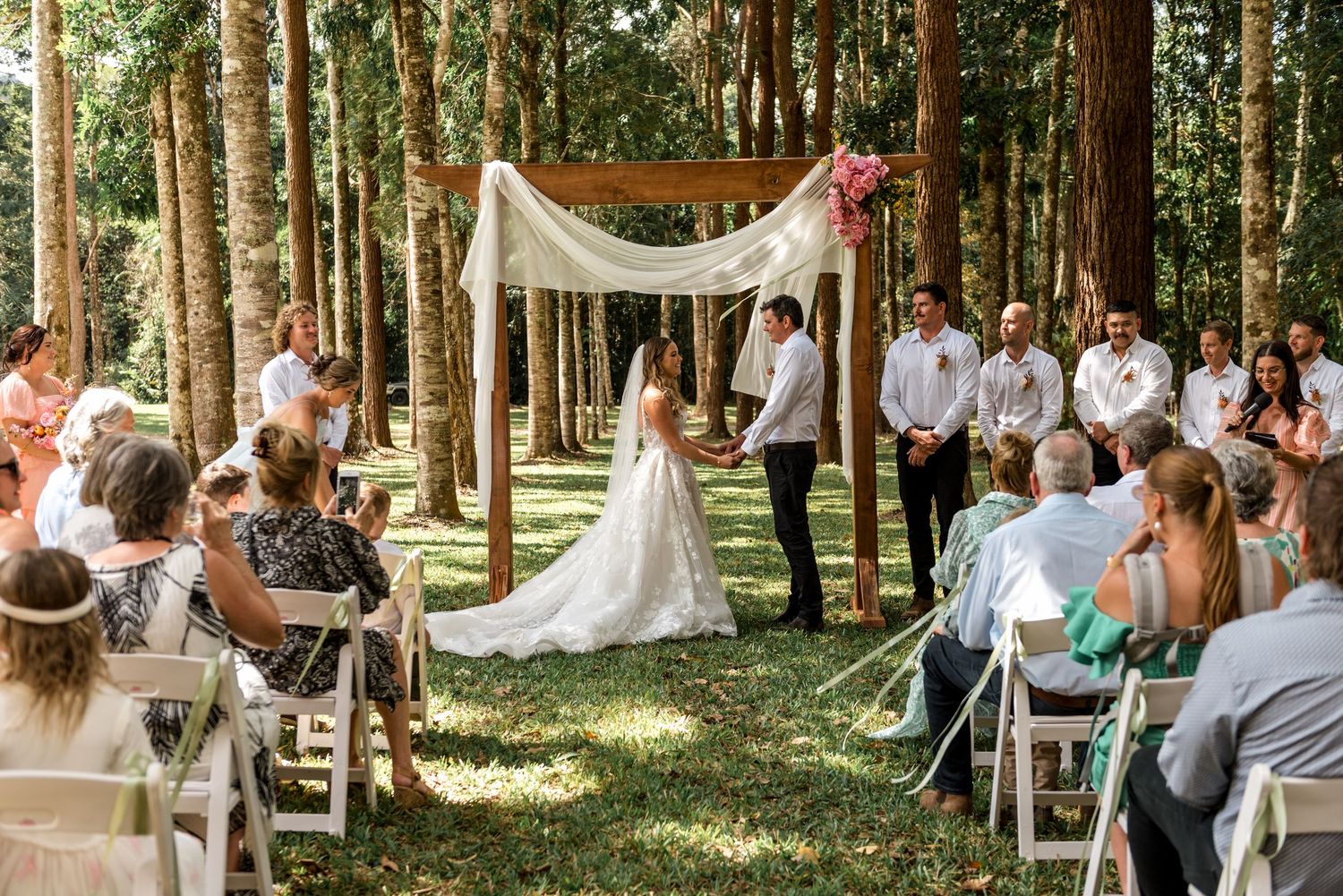Bride and groom exchanging vows under a draped wooden arch in a lush forest ceremony.