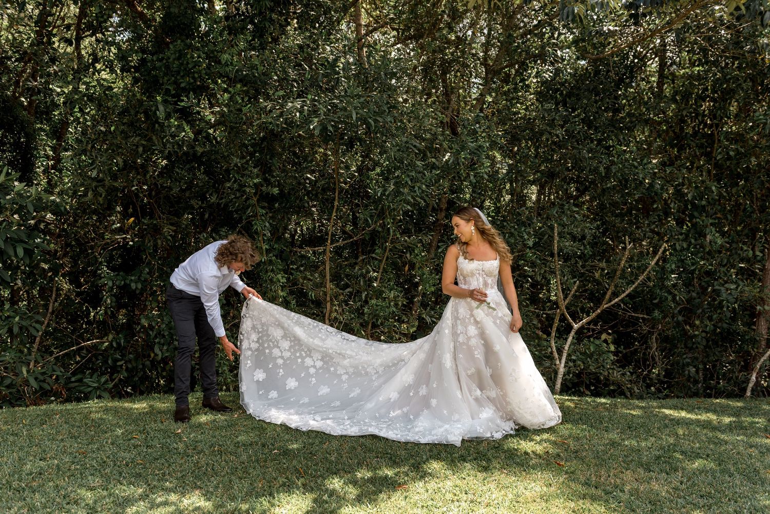 Groom adjusts elegant lace wedding dress train outdoors surrounded by dense green foliage.