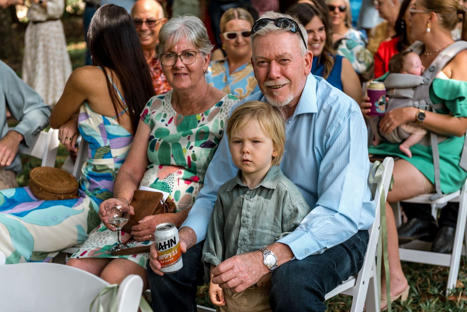 Grandparents and young boy pose together as wedding guests seated in white chairs outdoors.