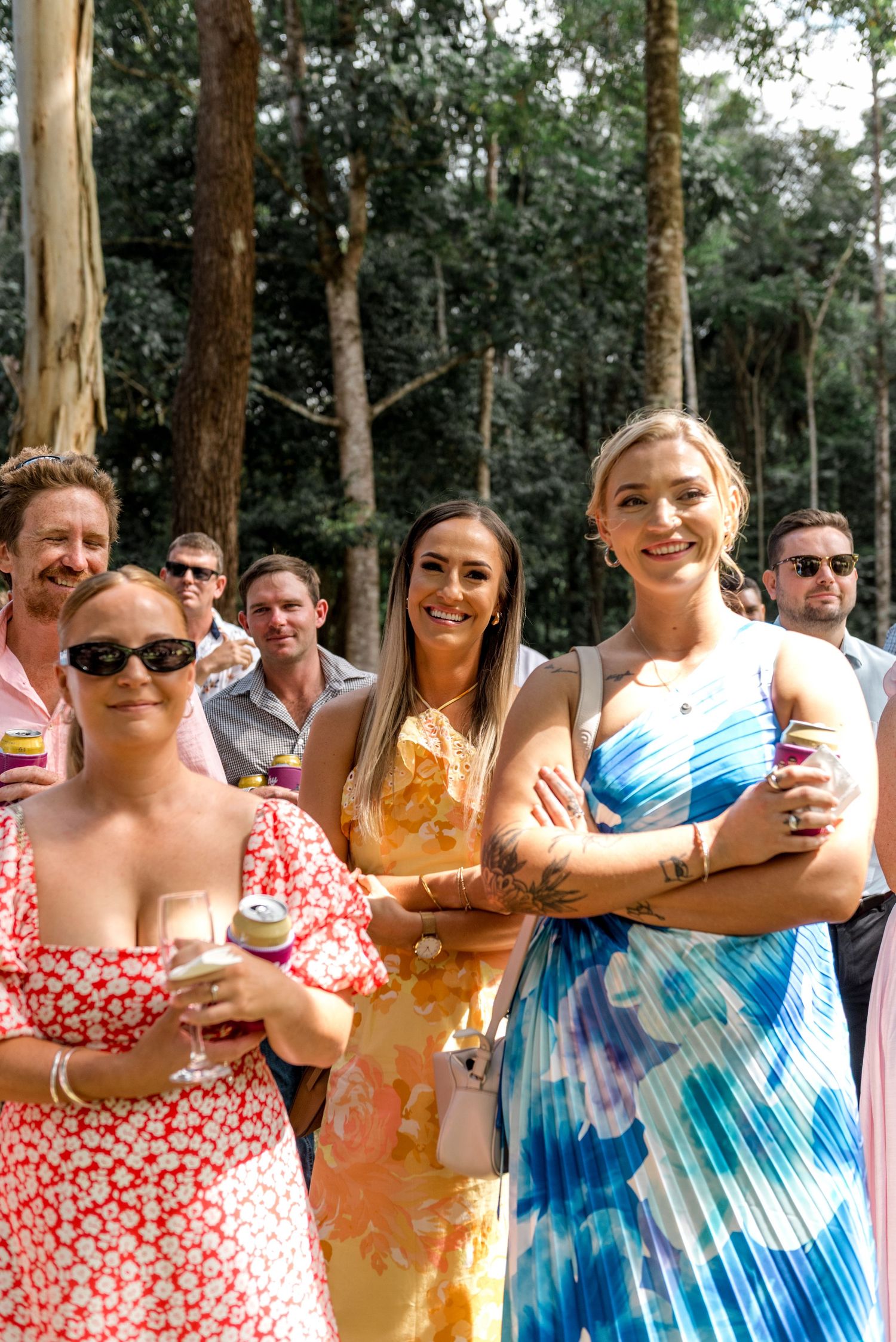 Smiling wedding guests in colorful floral dresses watch outdoor ceremony among tall trees.