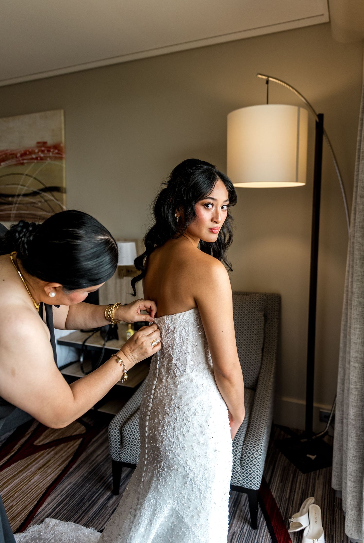 Woman helping bride button up elegant white lace wedding gown in a hotel room.