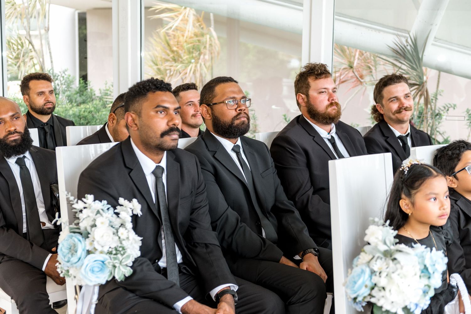 Male wedding guests in black suits seated at ceremony, with young flower girl holding blue bouquet.
