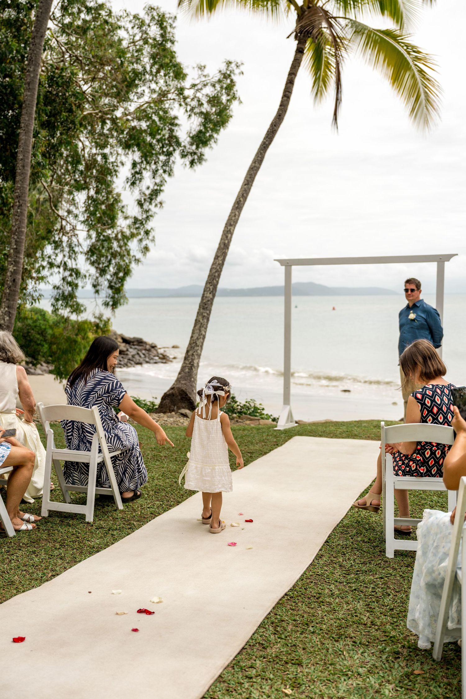 Flower girl walks down wedding aisle with ocean backdrop and white ceremony arch at Little Cove, Port Douglas.