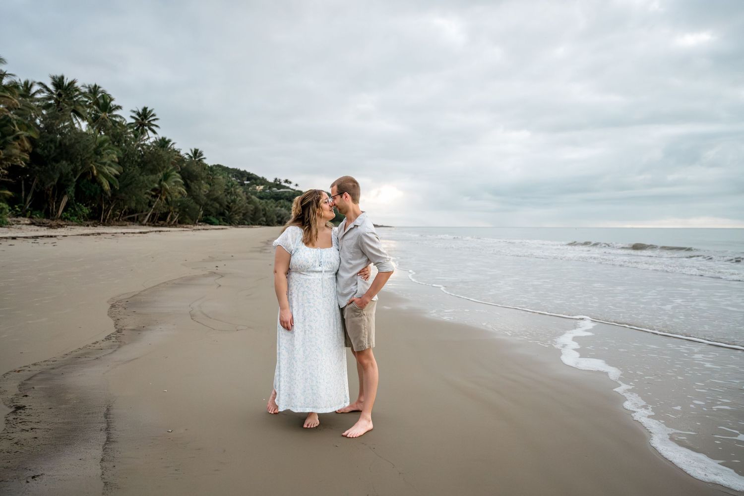 Romantic couple standing close together on a sandy beach with palm trees and cloudy sky at Four Mile Beach, Port Douglas.