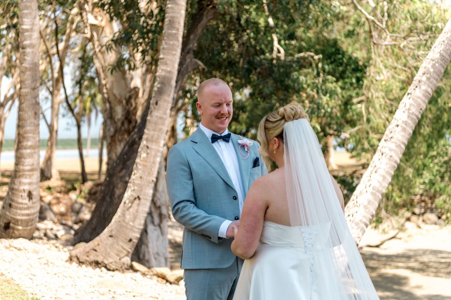 Emotional father seeing bride daughter for first time among palm trees before beach wedding ceremony.