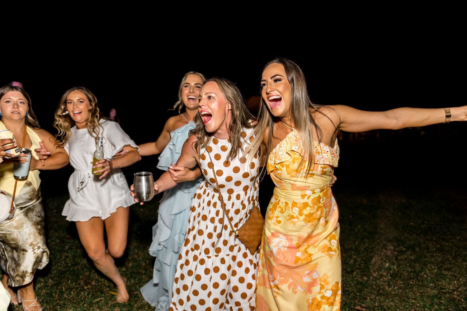 Joyful women in floral dresses dancing and laughing at outdoor nighttime wedding party.