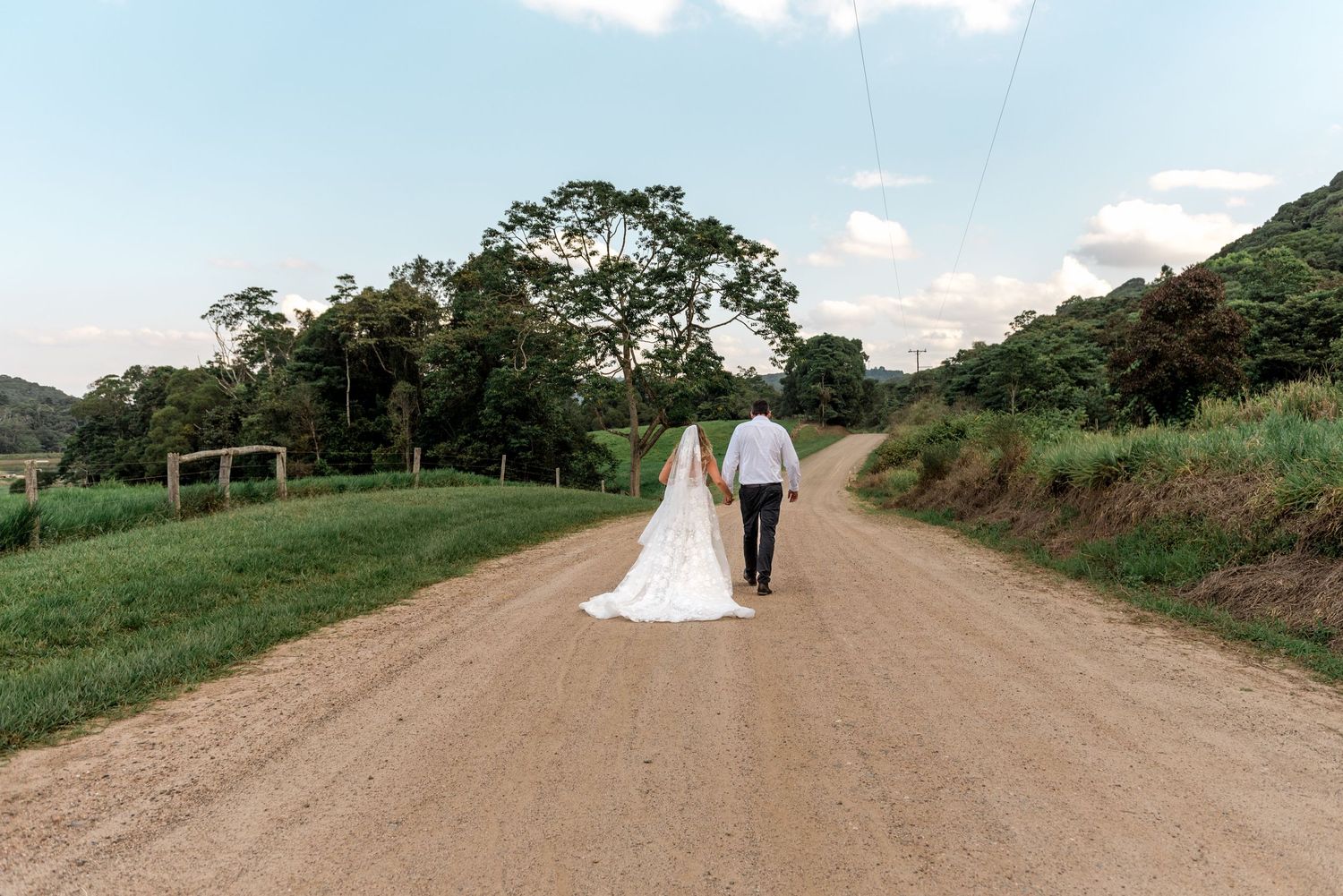 Bride and groom walk hand in hand down a wide gravel country road surrounded by lush green scenery.