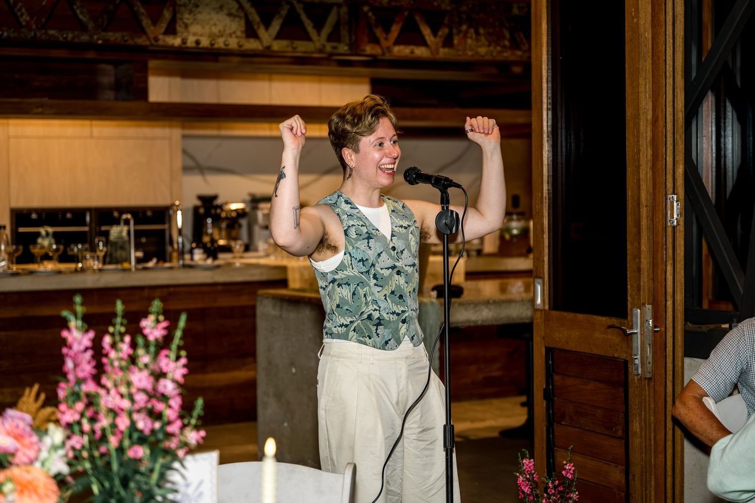 Woman enthusiastically giving a speech at a microphone inside a rustic venue with floral decor.