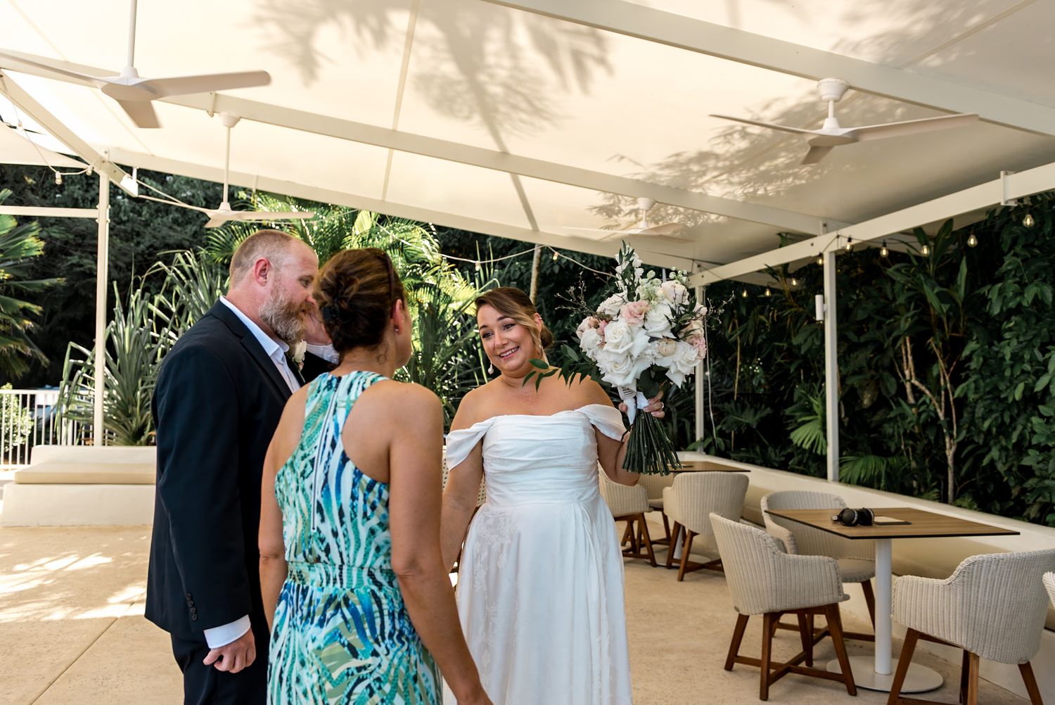 Bride in off-shoulder gown holds white orchid bouquet while chatting with guests under an outdoor wedding canopy.