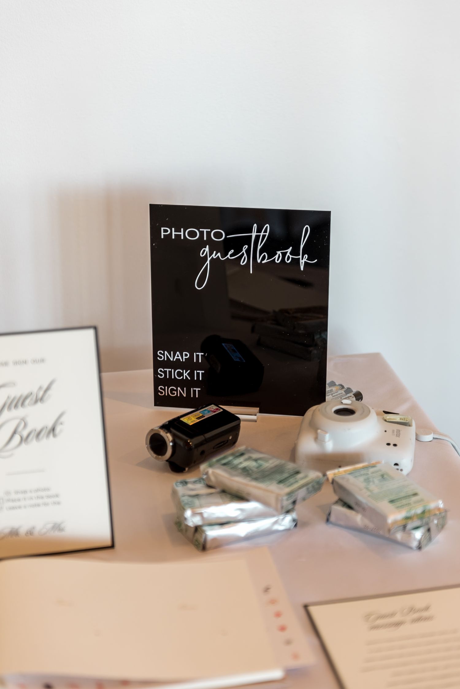 Wedding photo guestbook station with Instax camera, film packs, video camera, and snap-stick-sign instructions on display.