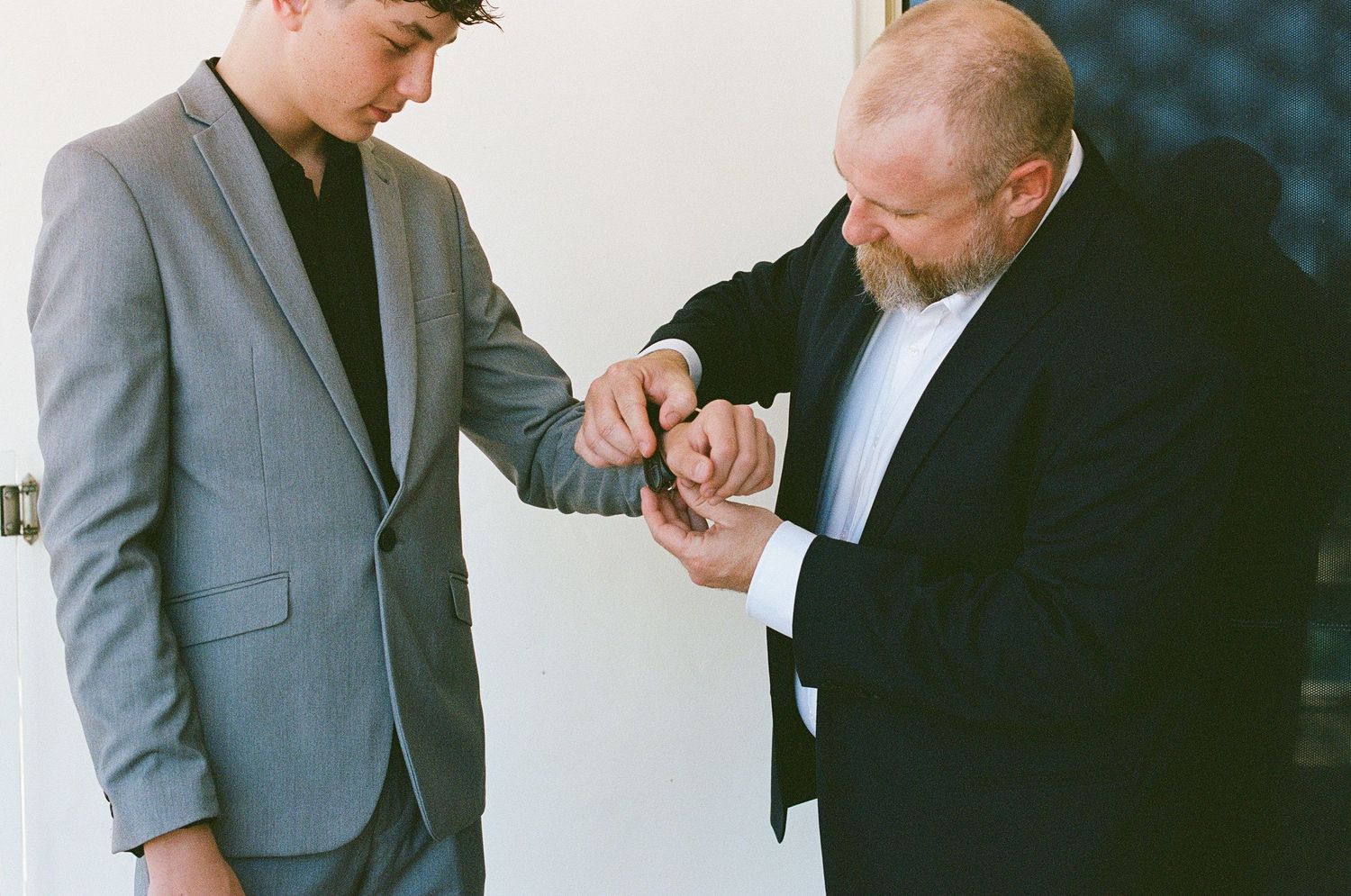 Man in gray suit getting cufflinks fastened by older man in black suit before wedding ceremony.