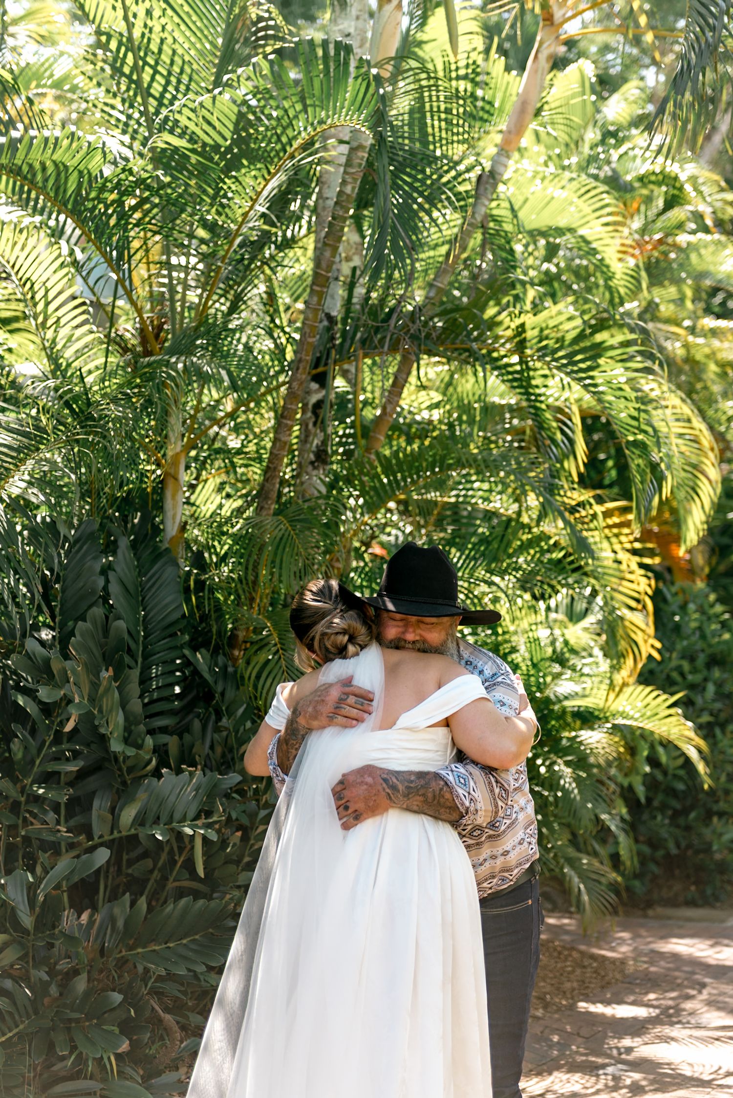 Bride and groom sharing a warm embrace surrounded by lush tropical palm trees in sunlit garden.